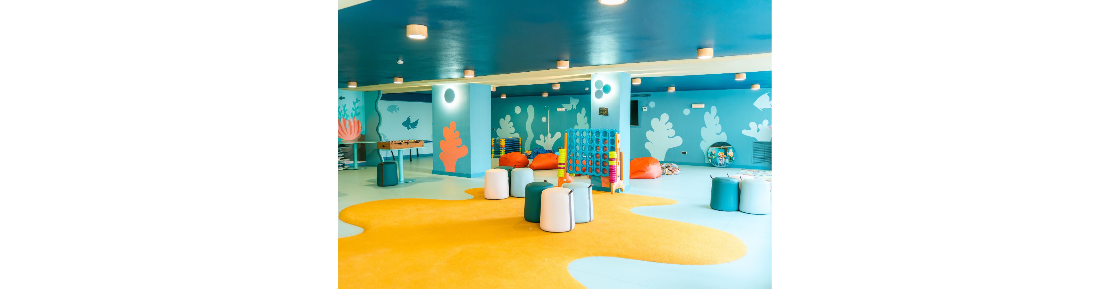 A bright indoor playroom features an ocean-themed design with blue walls and illustrated sea plants and fish. The space includes soft seating such as bean bags and round stools arranged on a large yellow rug. A giant connect four game and other play equipment are visible in the background, suggesting a fun area for children. Warm ceiling lights and playful colors create a cheerful, welcoming atmosphere.