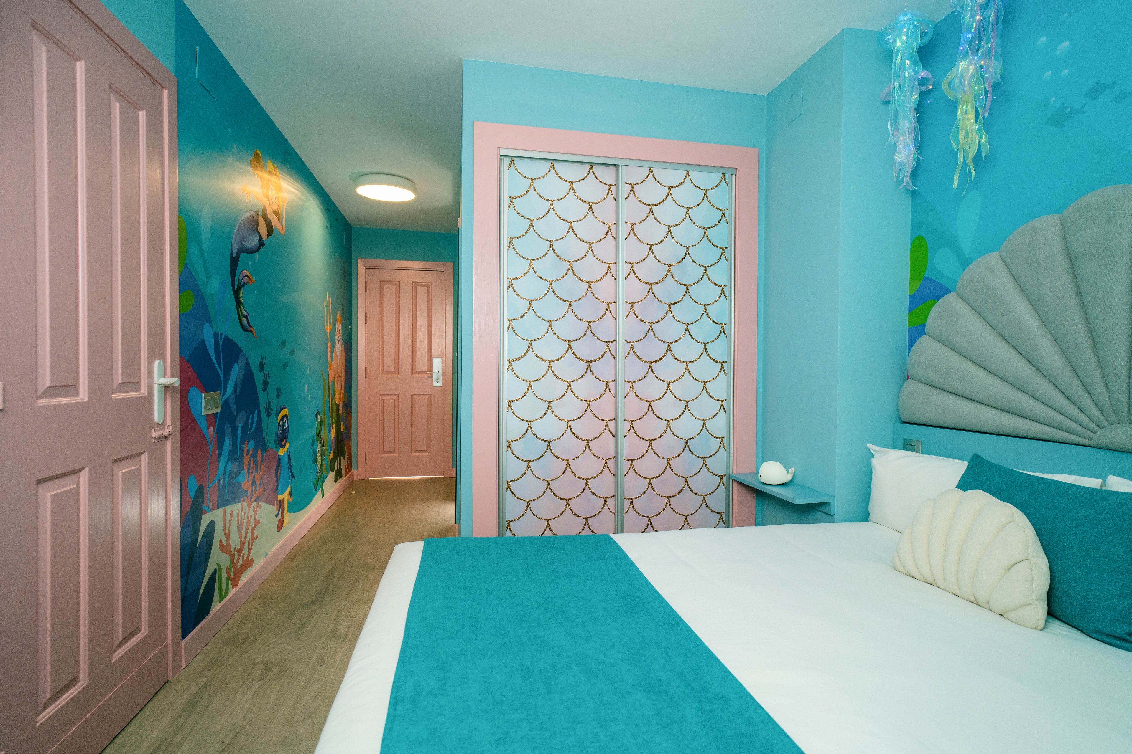 A vibrant bedroom features an undersea theme with turquoise walls and aquatic murals. The bed has teal bedding, shell-shaped pillows, and a scallop-inspired headboard. Pink doors and a wardrobe with fish-scale patterned panels add playful contrast. Jellyfish decorations hang from the ceiling, enhancing the whimsical underwater atmosphere.