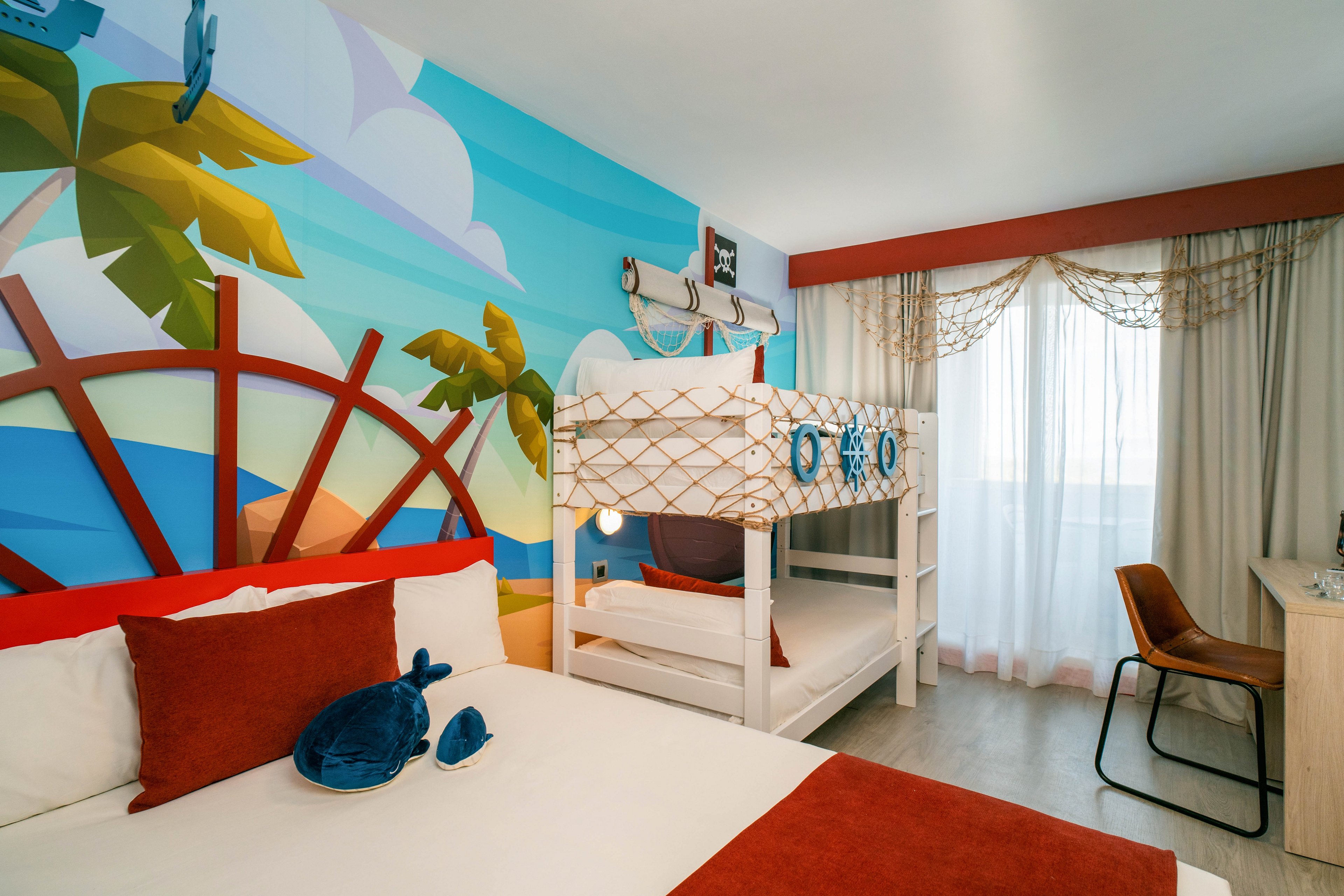 A bright family hotel room features a large bed in the foreground and a bunk bed decorated with nautical details. The wall is covered with a colorful mural showing a ship wheel, palm trees, and ocean scenery, creating a playful atmosphere. Sunlight filters through sheer curtains over a large window, illuminating the cozy seating area and desk. Red and white bedding with plush toys adds warmth and comfort to the child-friendly space.