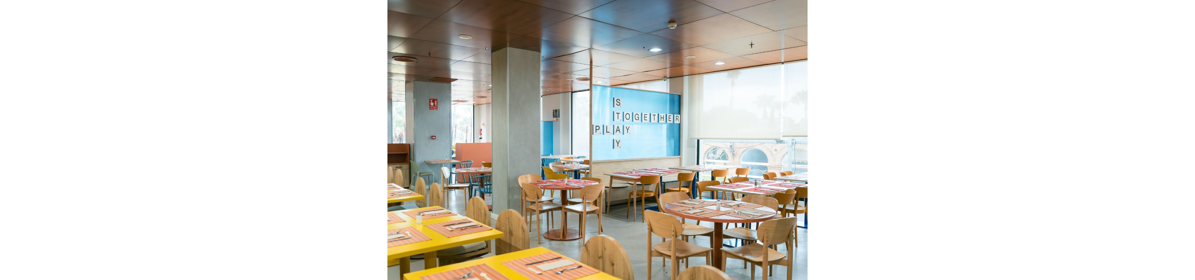 A spacious, modern cafeteria is filled with wooden tables and chairs neatly arranged in rows. Colorful placemats and table settings add warmth to the bright interior. Large windows let in natural light and reveal an outdoor view. On the back wall, oversized letter tiles spell out a playful message including the words "TOGETHER" and "PLAY".