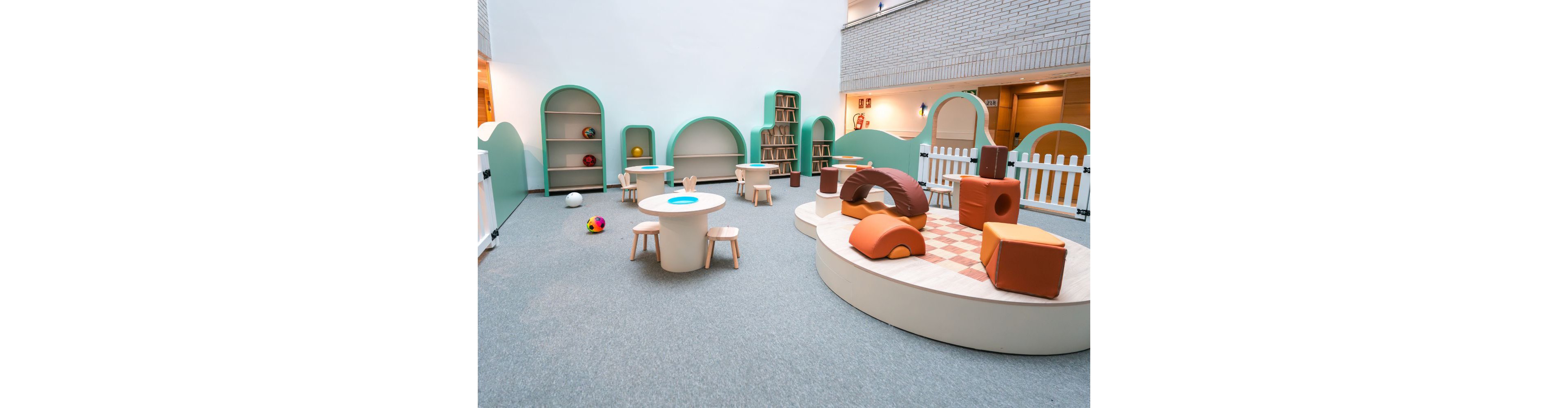 A bright indoor playroom features soft, colorful climbing blocks and low platforms designed for young children. Small round tables with matching stools sit on a carpeted floor, surrounded by curved shelves and play structures. A white picket fence and pastel green partitions define the play area within a larger building interior. Various toys, including balls and cubes, add playful accents to the minimalist, child-friendly space.