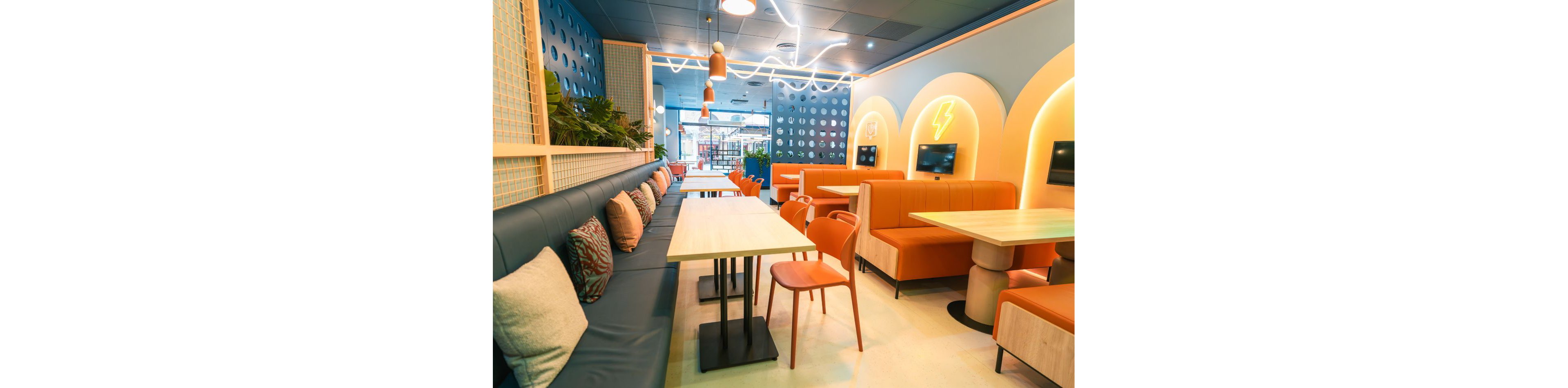 Bright contemporary cafe interior featuring long bench seating with cushions and individual booths. Light wooden tables and orange chairs create a playful contrast against blue walls and upholstery. Neon-style wall accents and soft strip lighting add a warm, inviting glow. Built-in screens and geometric decor elements give the space a modern, tech-friendly atmosphere.