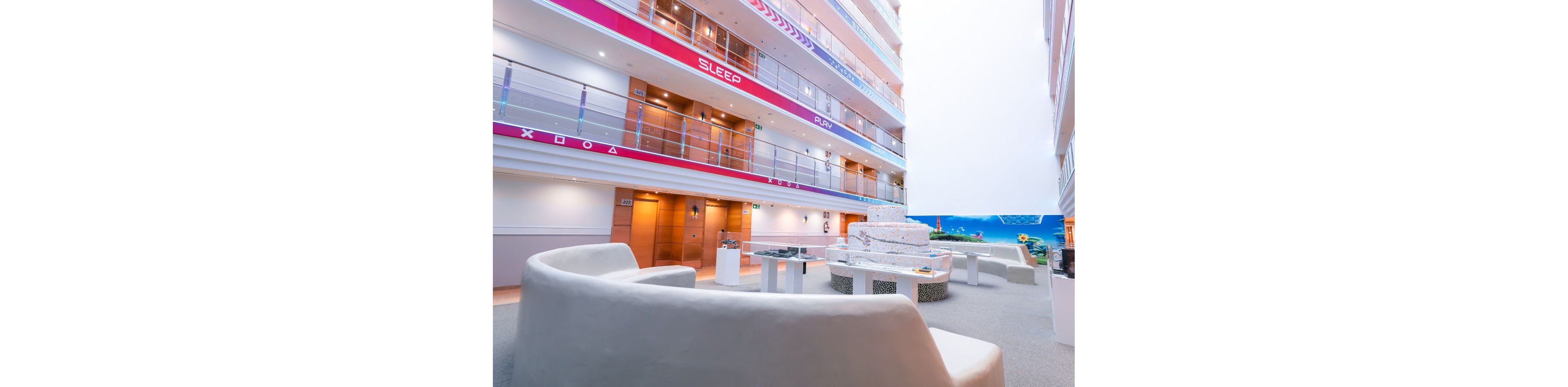 Bright contemporary lounge seating is arranged in the atrium of a cruise ship or large vessel. Curved white sofas and low tables create a relaxed social area on a carpeted floor. Multiple decks with glass railings rise above, featuring colorful bands with the words SLEEP and PLAY. Warm wooden doors line the corridor, and a vibrant ocean-themed mural adds color to the space. The overall atmosphere is open, airy, and inviting for passengers.