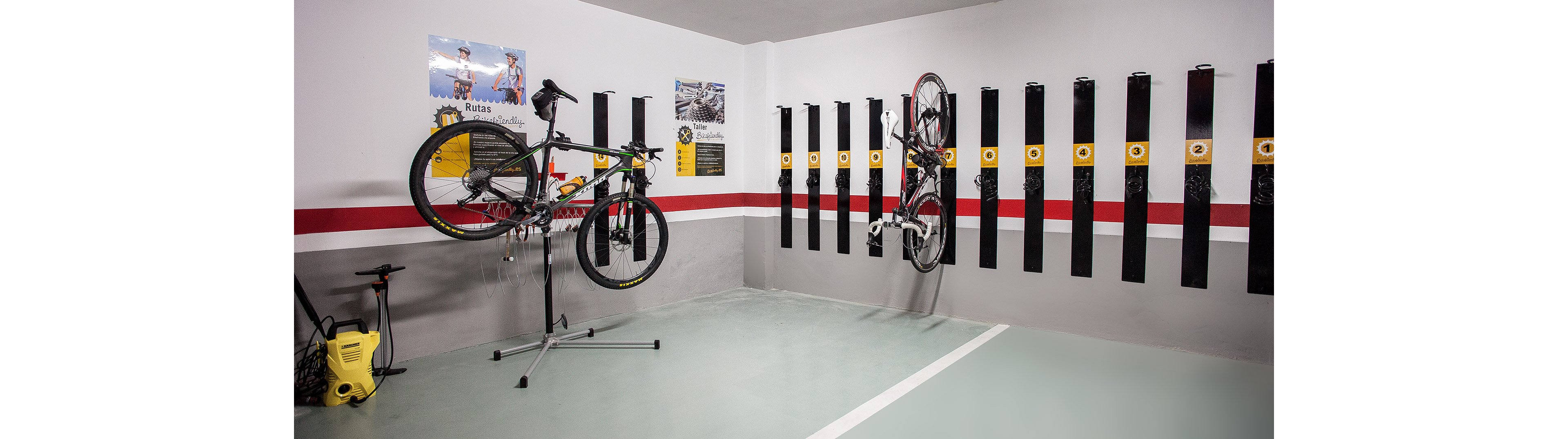 A clean indoor room is arranged for bicycle storage and maintenance. One bike is mounted on a repair stand while another hangs vertically on a wall rack. Multiple empty vertical bike racks line the wall, and cycling posters decorate the space. The floor and walls are painted in neutral tones with a bold red stripe for visual contrast.