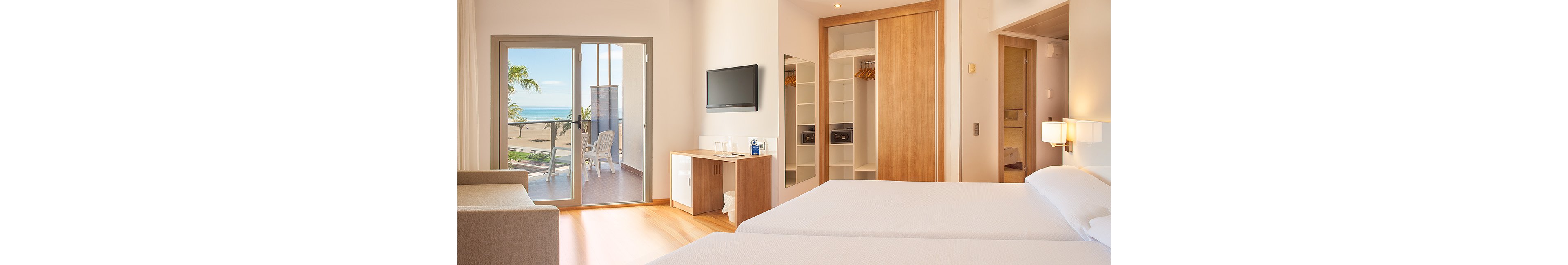 A modern hotel room features two neatly made beds and light wood furnishings. Large sliding glass doors open onto a balcony with chairs and a view of the ocean and palm trees. A flat-screen television is mounted on the wall above a small desk area. Open closets with shelves and safes, along with soft neutral colors, create a clean and relaxing atmosphere.