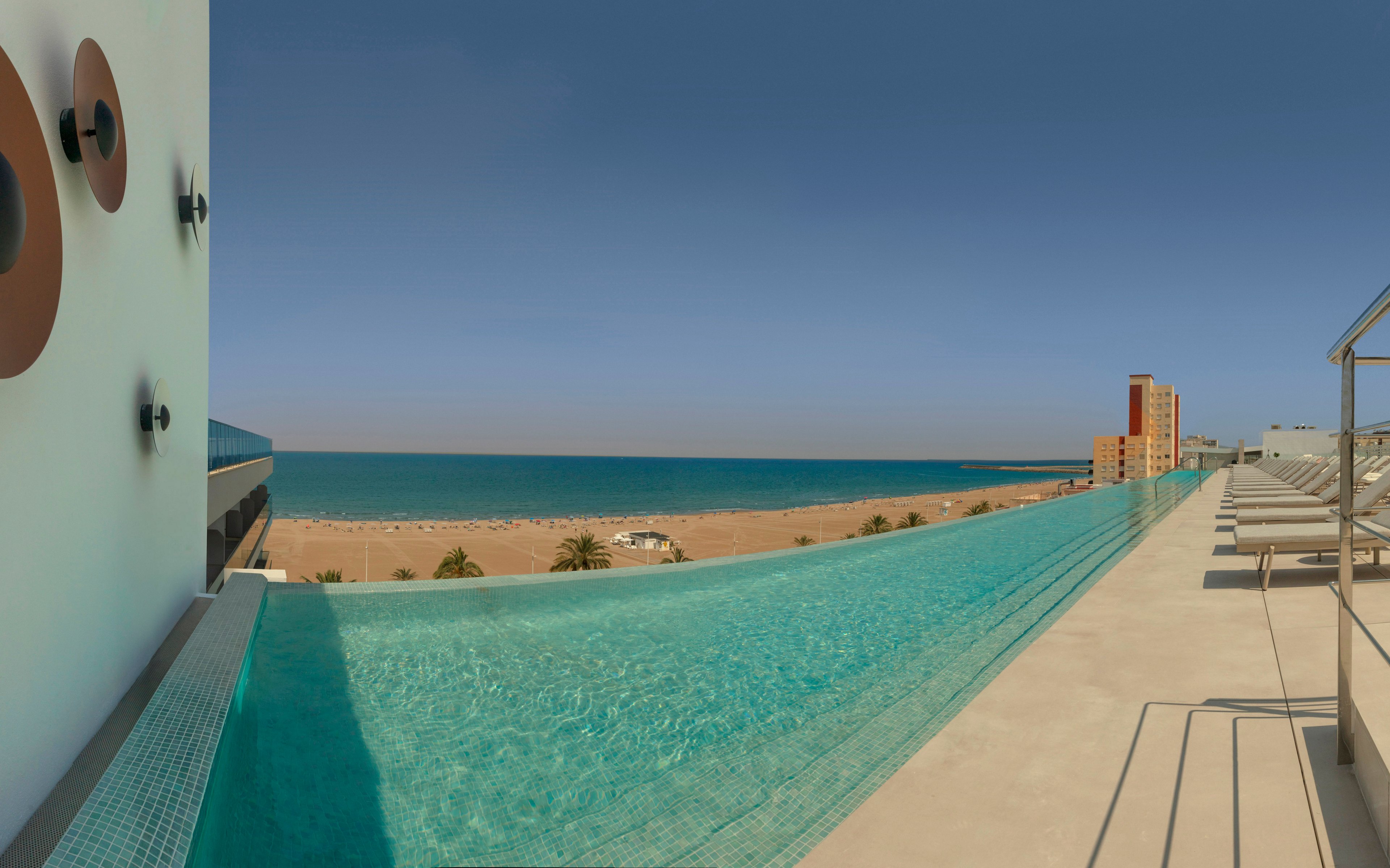 A young woman and man relax in a rooftop infinity pool overlooking a sandy beach and calm sea. The woman in a turquoise bikini sits at the pool edge while the man swims nearby. Modern wall lights and sleek railings frame the contemporary hotel terrace. Sun loungers line the deck, and tall coastal buildings are visible in the distance under a clear blue sky.