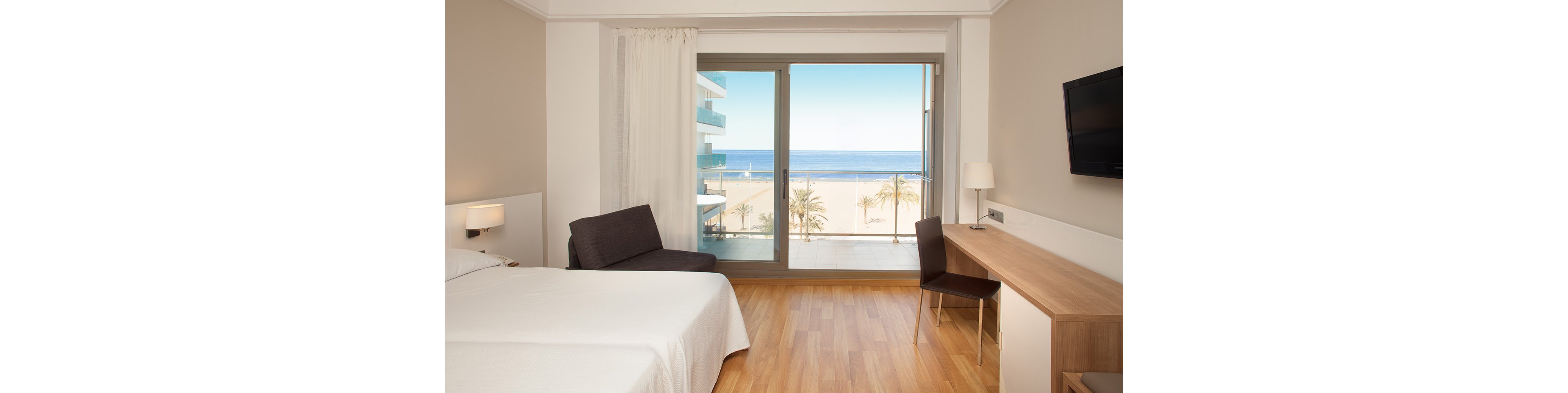 A modern hotel room features a large bed, wooden floors, and minimalist decor. A woman in a red dress relaxes on a chair while a man stands on the balcony looking out at the sea. Floor-to-ceiling glass doors open to a view of the beach, palm trees, and ocean horizon. A long wooden desk and chair line one wall, emphasizing the room’s spacious and bright atmosphere.
