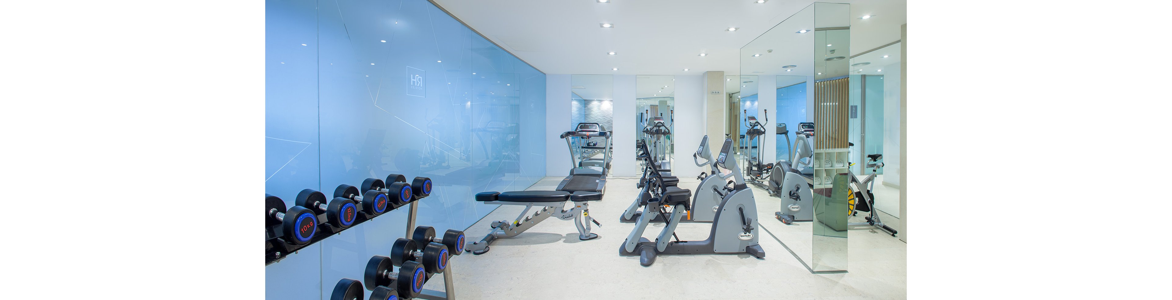 A bright, modern gym features a row of dumbbells, a weight bench, and cardio machines. The space has light-colored flooring, blue glass walls, and large mirrors that visually expand the room. Recessed ceiling lights create an evenly lit, clean atmosphere. The gym appears empty and ready for guests to exercise.