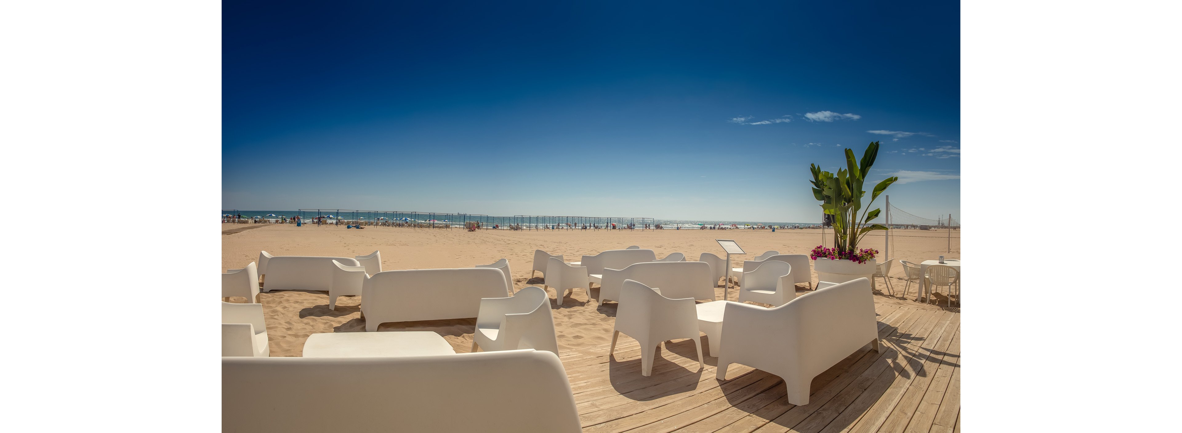 A row of modern white lounge chairs and sofas sits on a wooden deck overlooking a sandy beach. The scene is bright and sunny with a clear blue sky and calm sea in the background. Empty seating areas suggest a tranquil, relaxing seaside setting. A few potted plants add a touch of greenery to the minimalist outdoor design.