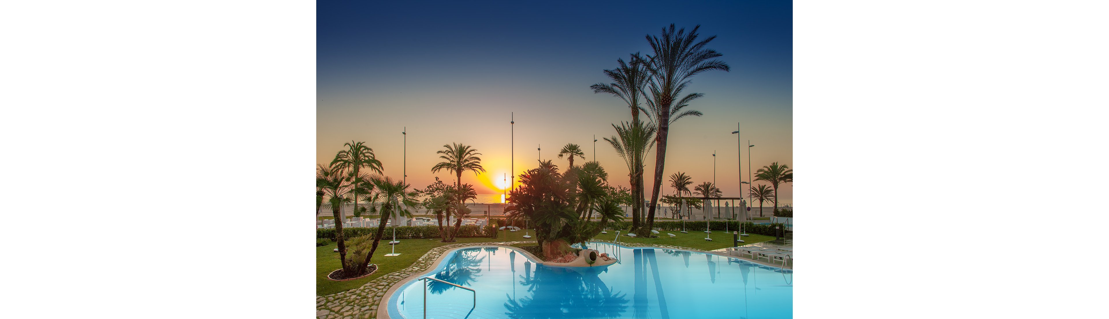 A tranquil outdoor swimming pool reflects the warm colors of a seaside sunset. Tall palm trees and lush greenery frame the water, creating a tropical resort atmosphere. Lounge chairs and umbrellas line the poolside, suggesting a relaxing vacation setting. The calm sea and clear sky in the background enhance the peaceful, luxurious mood.