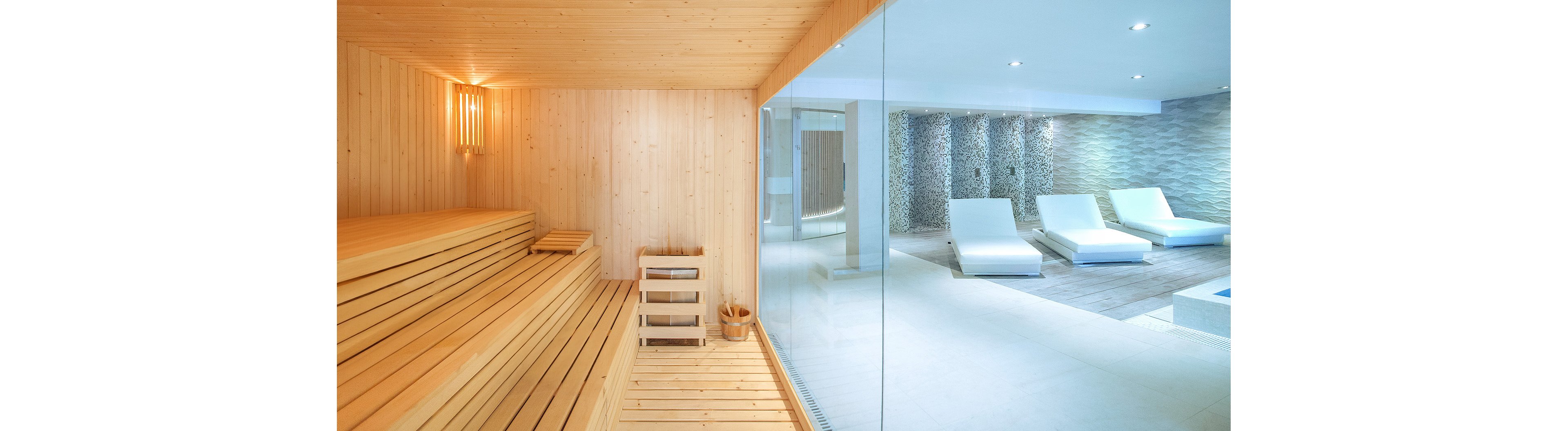 Several women relax in a contemporary spa that combines a wooden sauna and a bright lounge area. On the left, two women in swimwear recline on wooden benches inside a warm, light-wood sauna. A glass wall separates the sauna from the spacious relaxation room with white loungers and soft lighting. The overall atmosphere is calm, luxurious, and focused on wellness and self-care.
