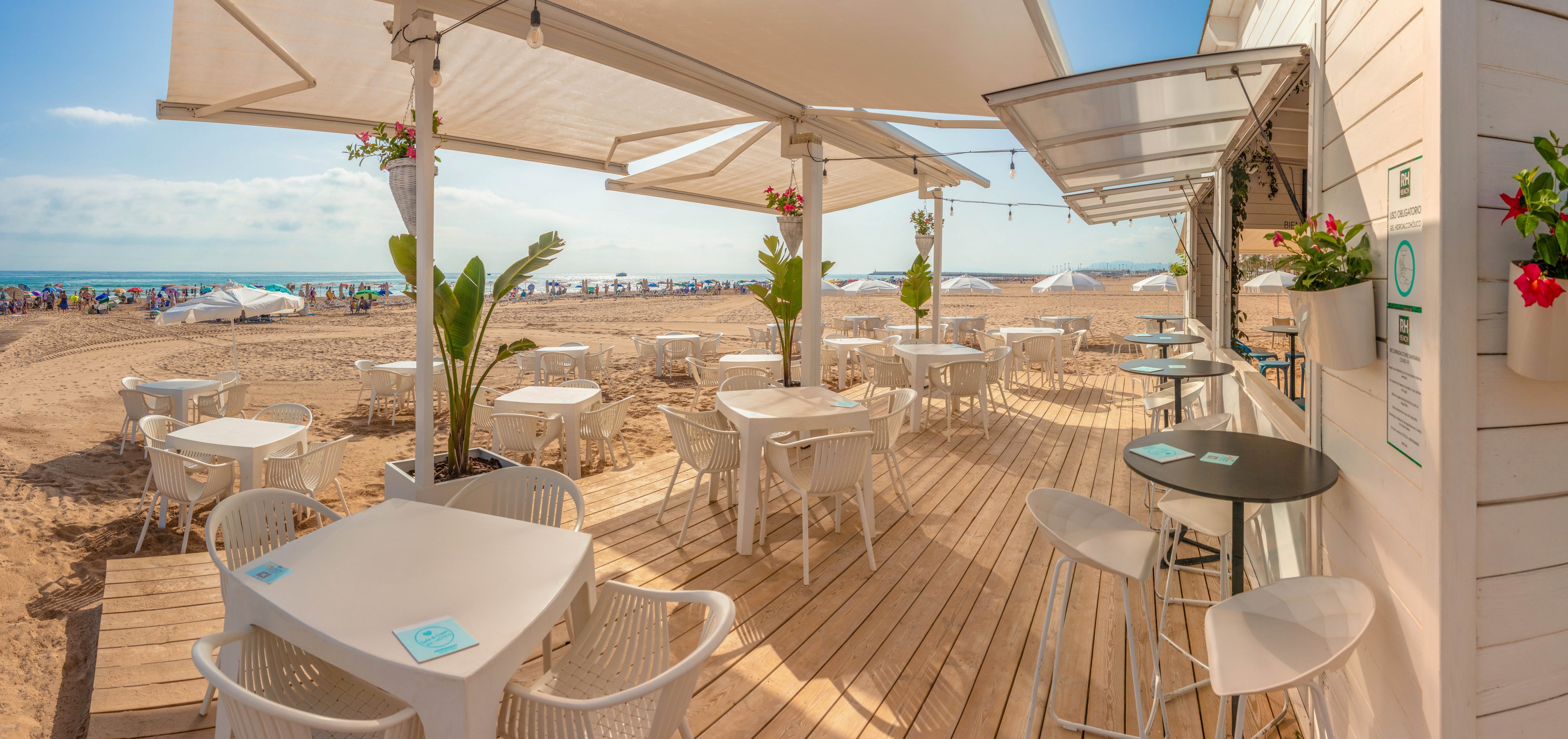 An open-air café terrace overlooks a sandy beach on a bright sunny day. White tables and chairs sit on a wooden deck beneath large awnings that provide shade. A few potted plants and red flowers add color to the light, neutral décor. In the background, beachgoers relax under umbrellas near the calm sea and blue sky.