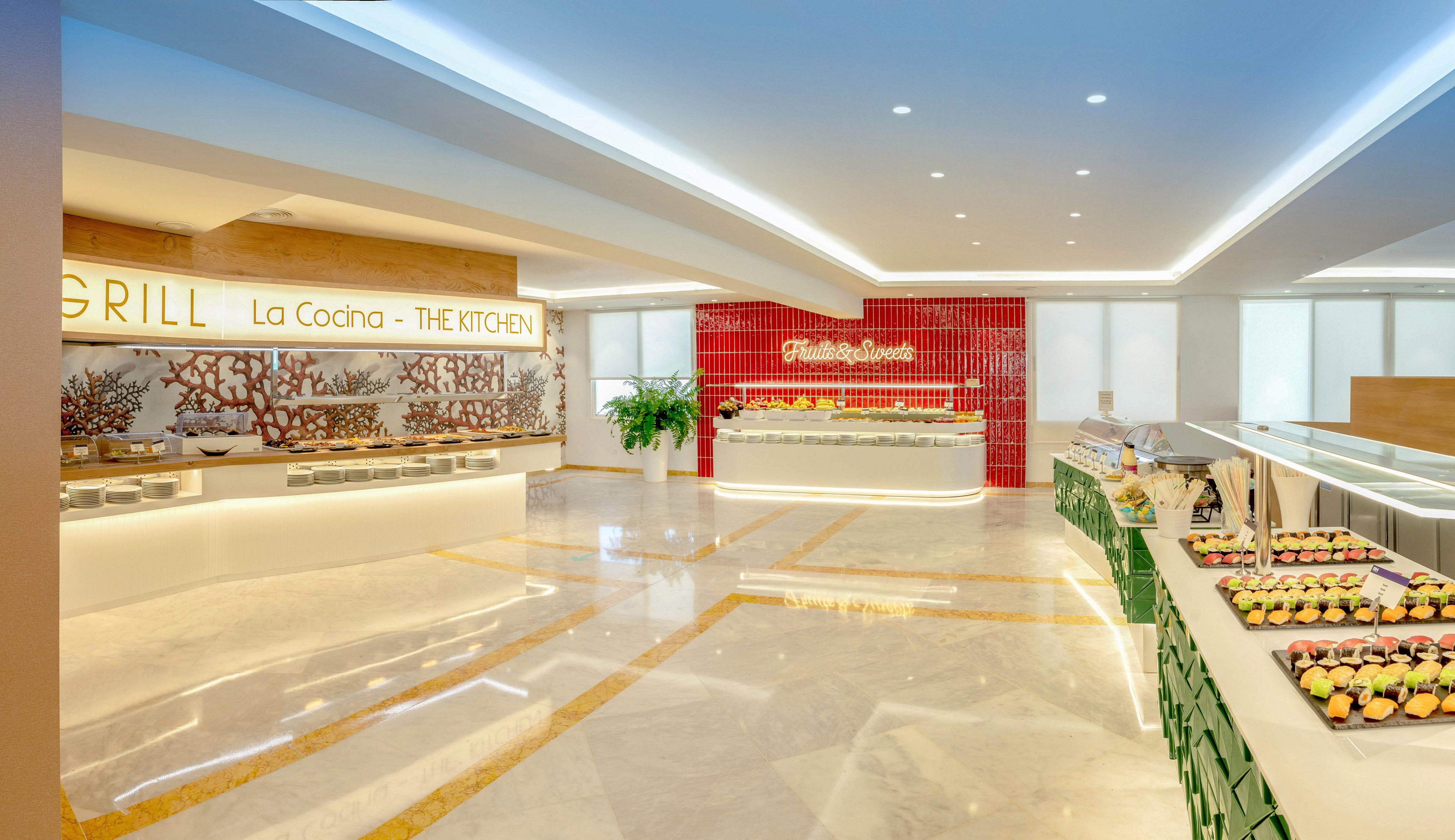 A spacious hotel buffet restaurant features multiple serving stations and a polished marble floor. On the left, a counter labeled "GRILL La Cocina - THE KITCHEN" offers food beneath warm lighting and decorative coral artwork. In the background, a red-tiled wall highlights another buffet area with illuminated script text. To the right, rows of neatly arranged desserts and pastries sit on green crates, adding color to the modern, minimalist interior. The scene is empty of people, emphasizing the clean, organized presentation.