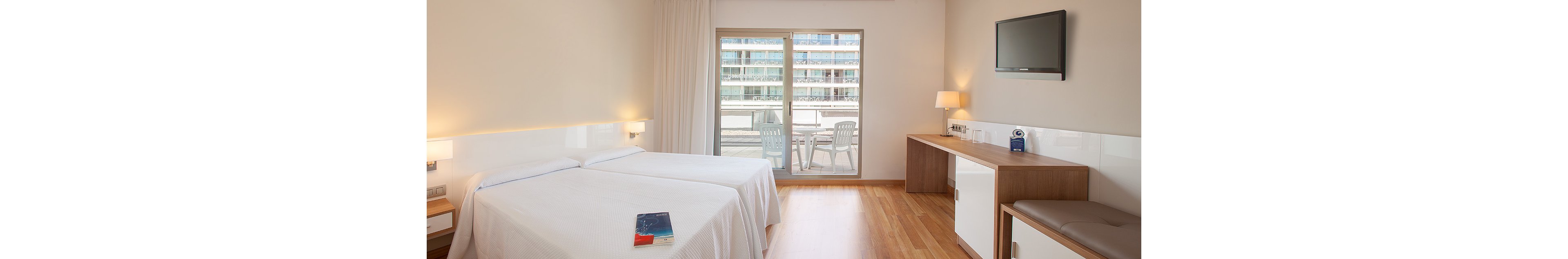 A bright hotel room features two neatly made single beds with white linens. Large glass doors open to a balcony overlooking neighboring buildings. A wall-mounted flat-screen TV faces the beds above a long wooden desk with a small refrigerator and chair. The room has light wood flooring, neutral walls, and soft lighting, creating a clean and minimalist atmosphere.
