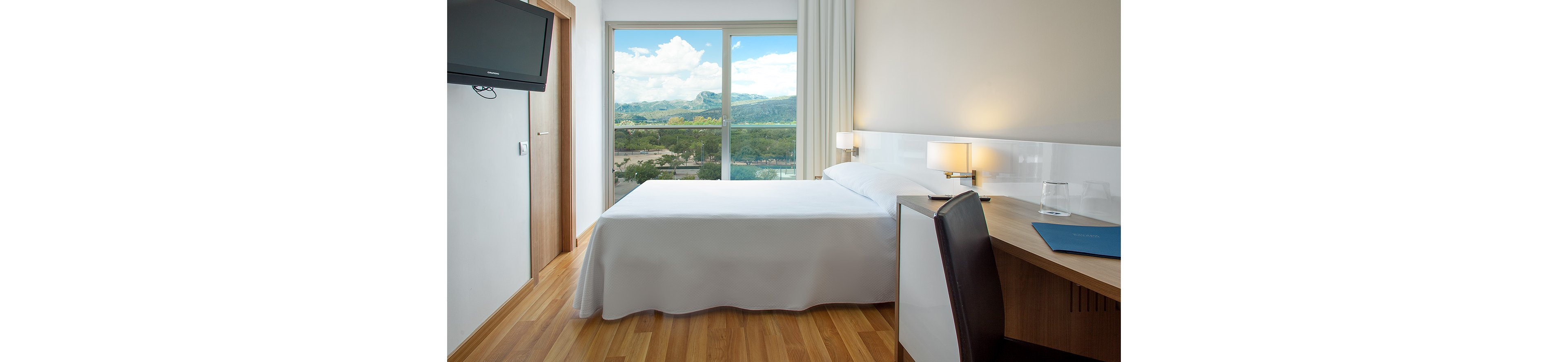 A modern hotel room features a neatly made double bed with white linens. Large sliding glass doors open to a scenic view of green hills and a blue sky with clouds. A wooden desk with a chair, lamp, and blue folder sits beside the bed, while a wall-mounted television hangs opposite. The room has light wood flooring and neutral walls, creating a clean and minimalist atmosphere.