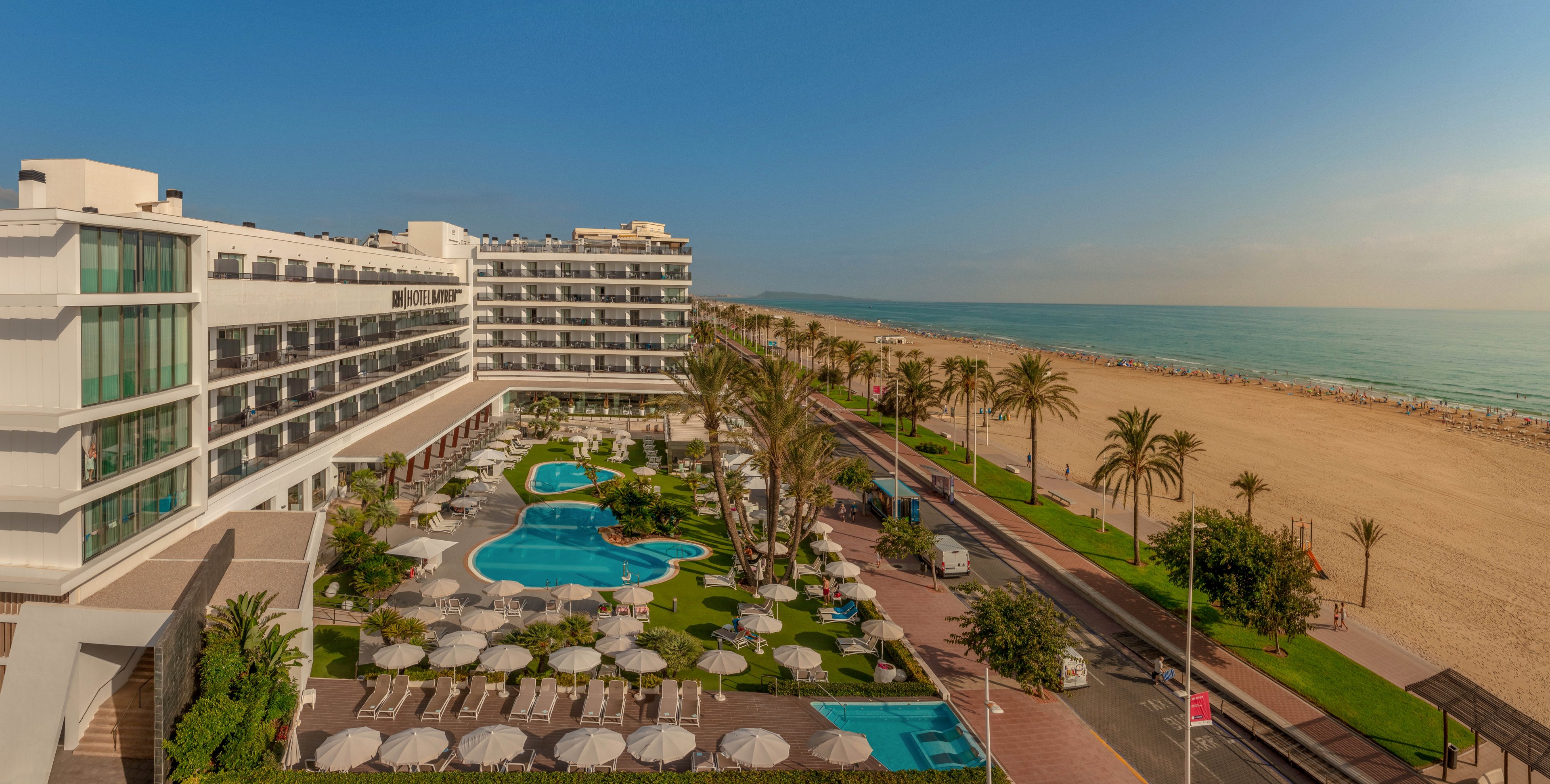 A large beachfront resort overlooks a spacious sandy beach and calm sea. The hotel features multiple stories of rooms with balconies facing a landscaped pool area. Curved swimming pools, sun loungers, and umbrellas are arranged neatly among palm trees and green lawns. A promenade with a few people walking separates the resort from the wide shoreline under a clear blue sky.