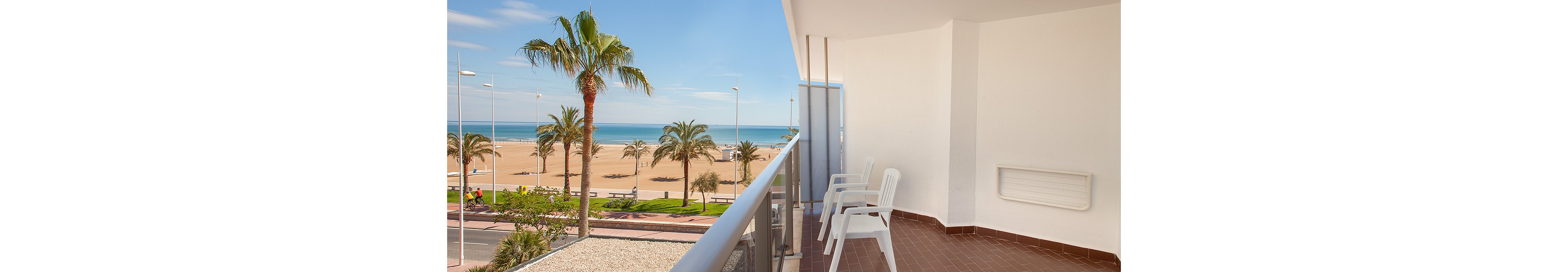 A spacious hotel balcony with white chairs overlooks a wide sandy beach and calm blue sea. Palm trees line the promenade between the building and the shoreline. The sky is clear with a few light clouds, creating a bright and inviting coastal scene. The modern balcony railing and tiled floor add a clean, contemporary feel to the seaside view.