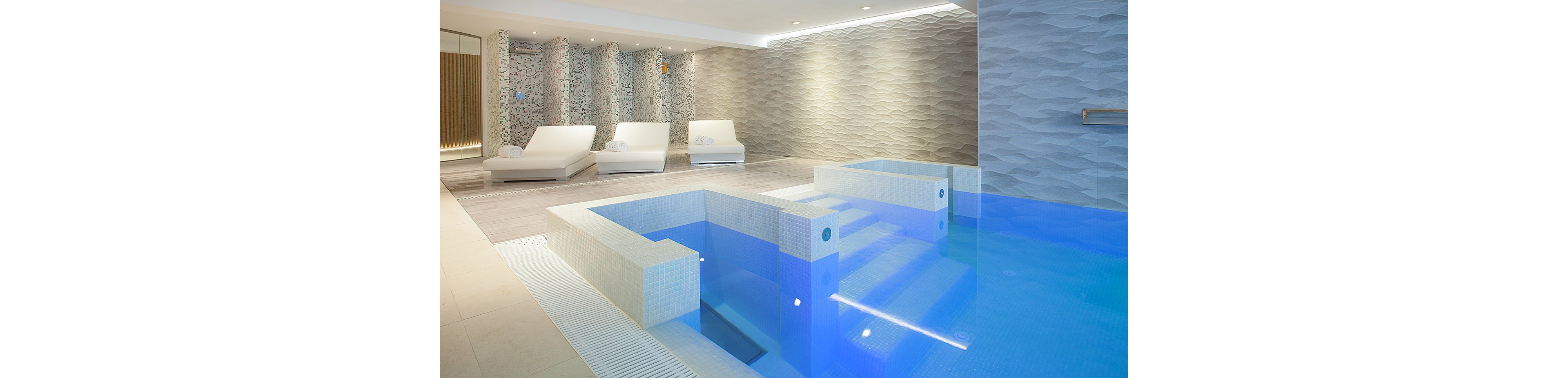 A contemporary indoor spa pool features clean white tiling and clear blue water. Built-in steps and submerged seating create a geometric layout within the pool. On the left side, several cushioned white loungers sit against a textured accent wall. Soft, indirect lighting and neutral tones give the space a calm, luxurious atmosphere.