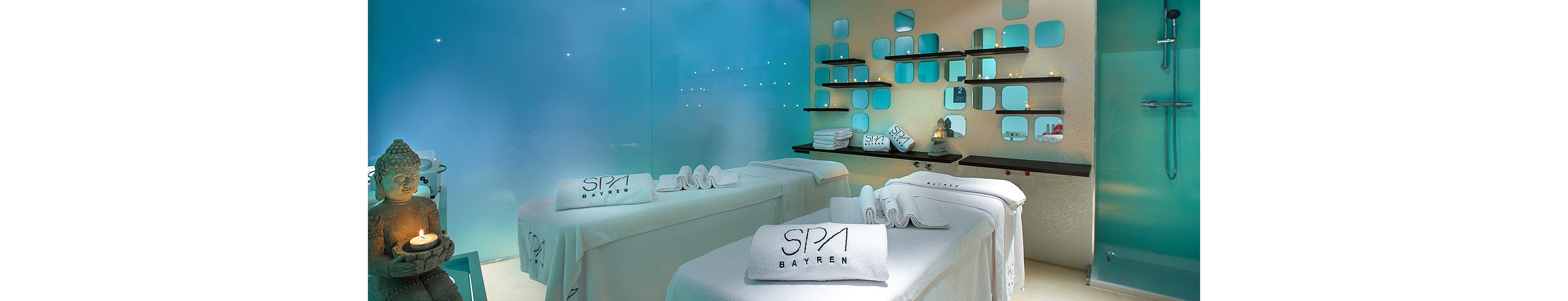 Contemporary spa treatment room with two massage tables covered in white linens. Soft blue lighting and frosted glass walls create a tranquil, modern atmosphere. Shelves with neatly arranged towels and products line the back wall, while a serene Buddha statue glows warmly in one corner. The word "SPA" is clearly visible on folded towels, emphasizing the wellness setting.