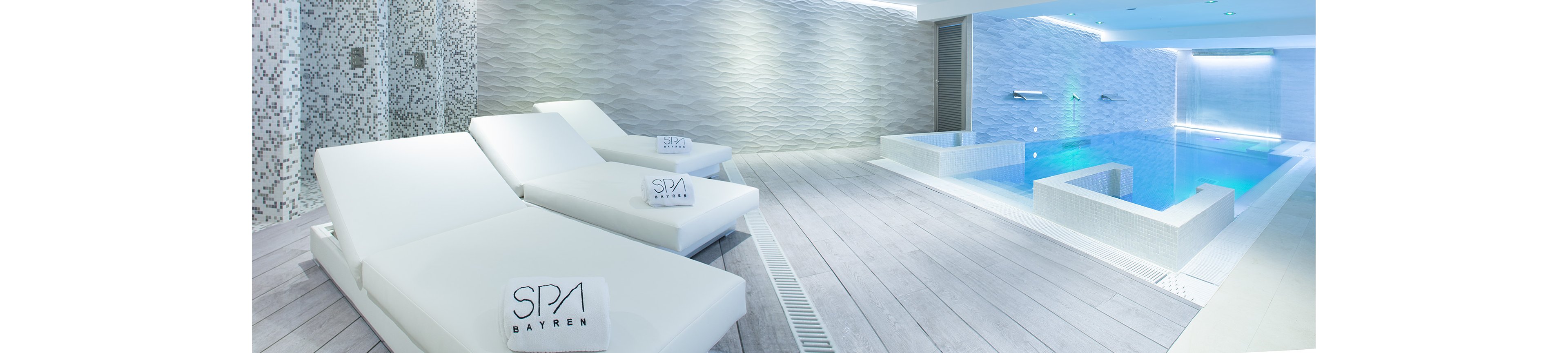 A contemporary indoor spa features sleek white loungers beside a tranquil pool. The space is finished with light wood-style flooring and textured stone walls. Soft blue lighting illuminates the water, creating a calm and luxurious atmosphere. Towels labeled "SPA" rest on the loungers, reinforcing the wellness setting.