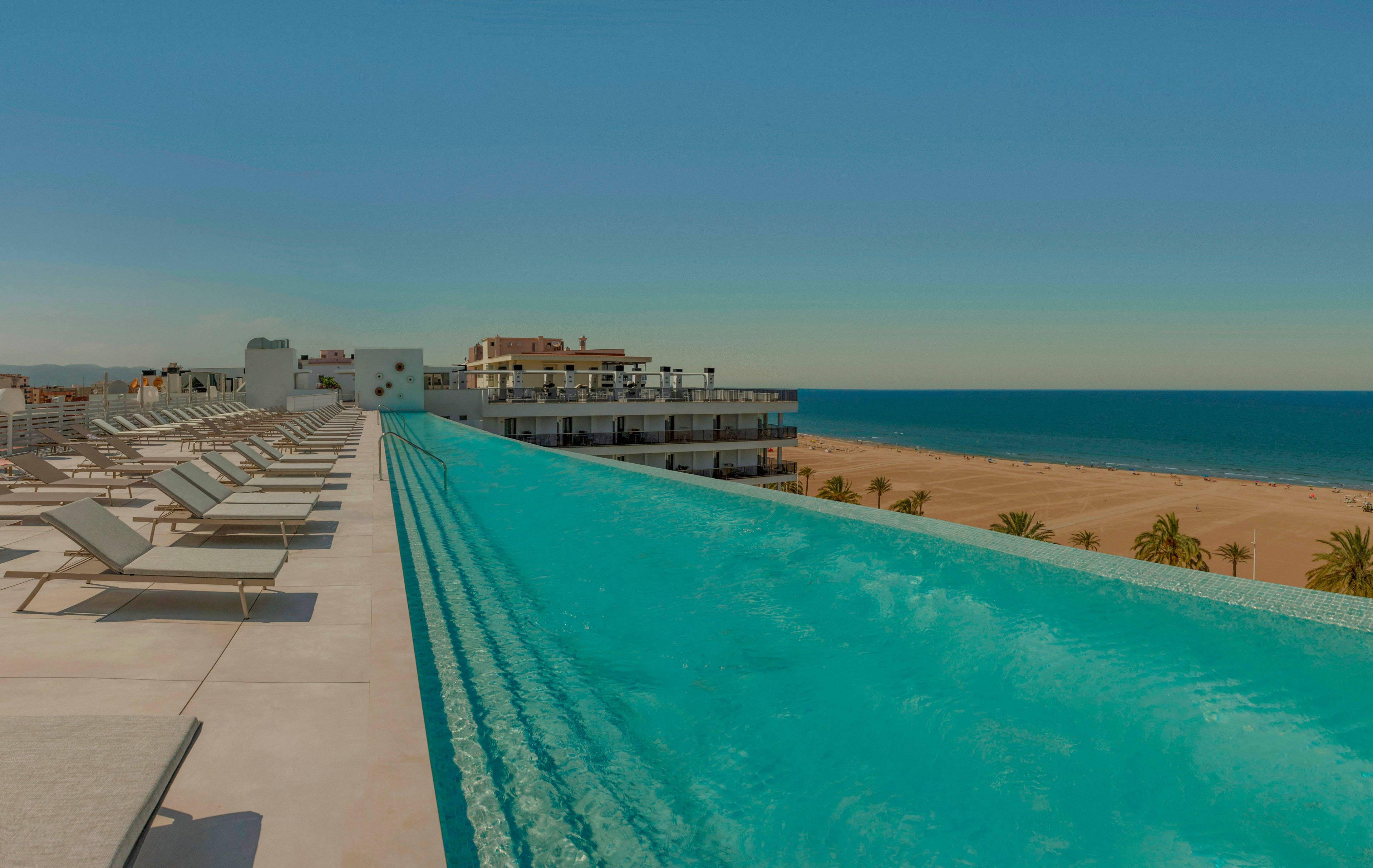 A swimmer glides through a long rooftop infinity pool with clear turquoise water. White lounge chairs line the pool deck under a clear blue sky. In the background, modern beachfront buildings and a wide sandy shoreline stretch along the coast. The calm sea and minimal design create a serene, luxurious vacation atmosphere.
