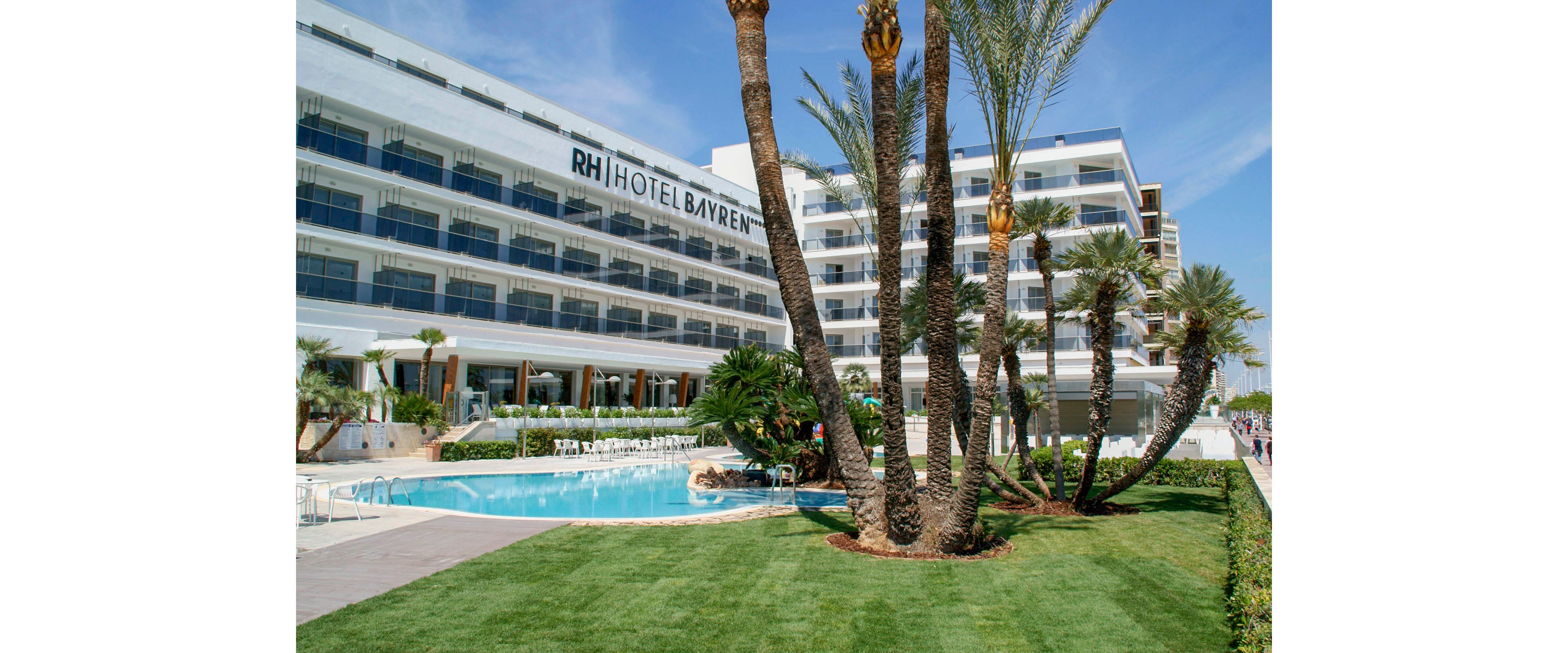 A large modern hotel building labeled RH Hotel Bayren stands beside an outdoor swimming pool. Tall palm trees and manicured green lawns frame the pool area, creating a resort-like atmosphere. White lounge chairs and umbrellas are arranged around the water, suggesting a relaxing holiday setting. The bright daylight and clear sky emphasize a warm coastal climate ideal for vacation stays.