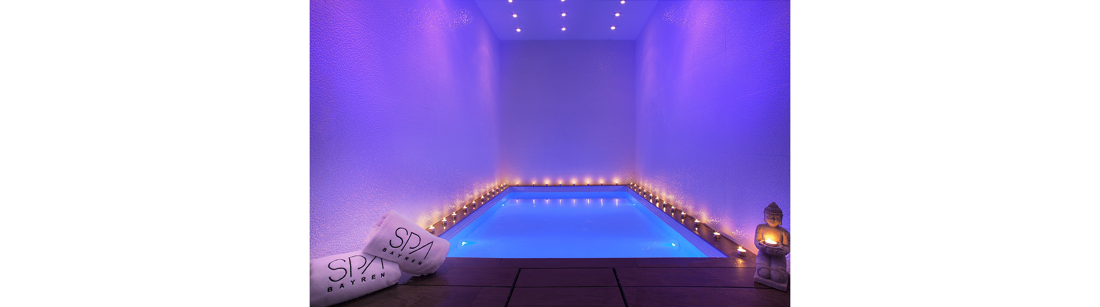 A serene indoor spa pool glows with soft blue lighting in a narrow, minimalist room. Rows of small candles line both sides of the water, creating a warm contrast against the cool illuminated pool. Rolled white towels with the text "SPA BAYREN" rest on a wooden deck in the foreground. A small Buddha statue with a candle adds a meditative touch to the relaxing atmosphere.