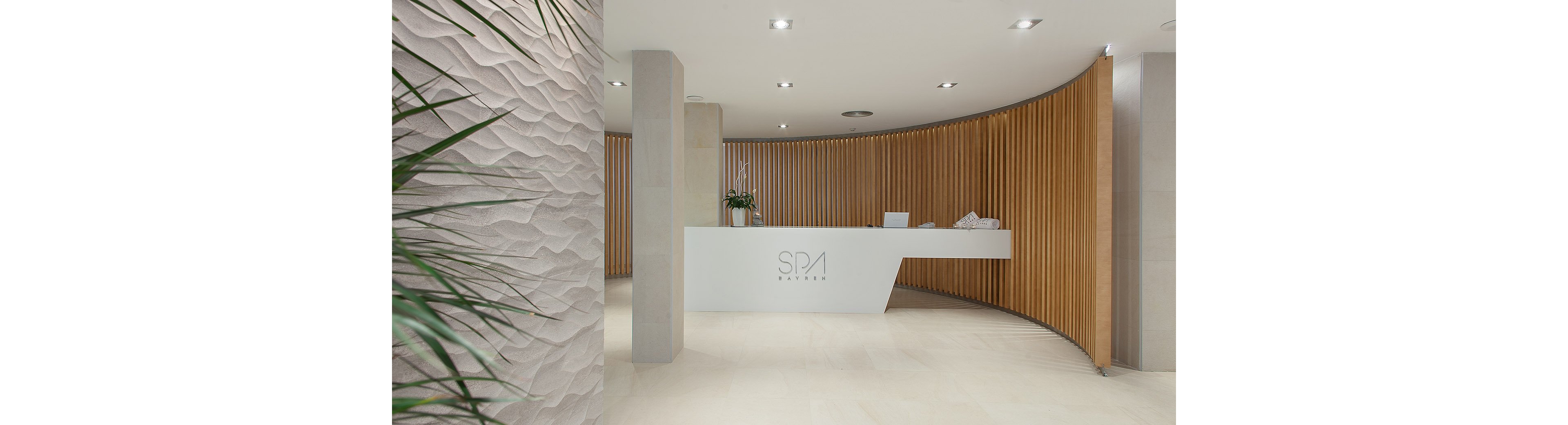 A minimalist spa reception area features a sleek white desk centered in front of a curved wooden slat wall. The space is bright and clean, with light-colored flooring and recessed ceiling lights. A textured wall with wave-like patterns appears on the left, adding visual interest. Green plants and neatly folded towels on the counter contribute to a calm, welcoming atmosphere.