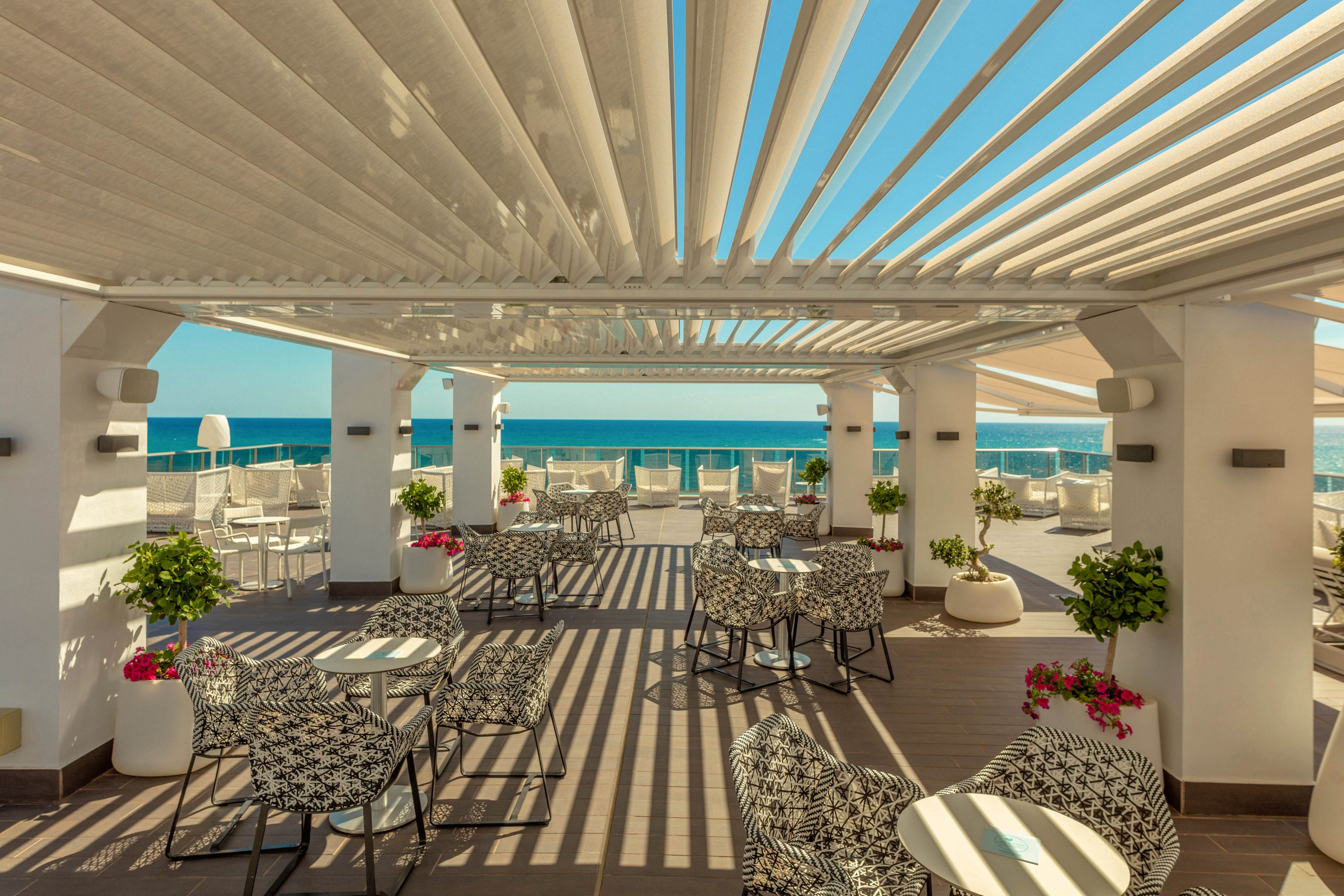 An open-air terrace café overlooks a bright blue ocean under a modern pergola roof. White columns and slatted shades create geometric shadows across the wooden deck flooring. Patterned black and white chairs surround small round tables, with potted plants and flowers adding color. The setting appears calm and inviting, ideal for relaxed outdoor dining or drinks by the sea.