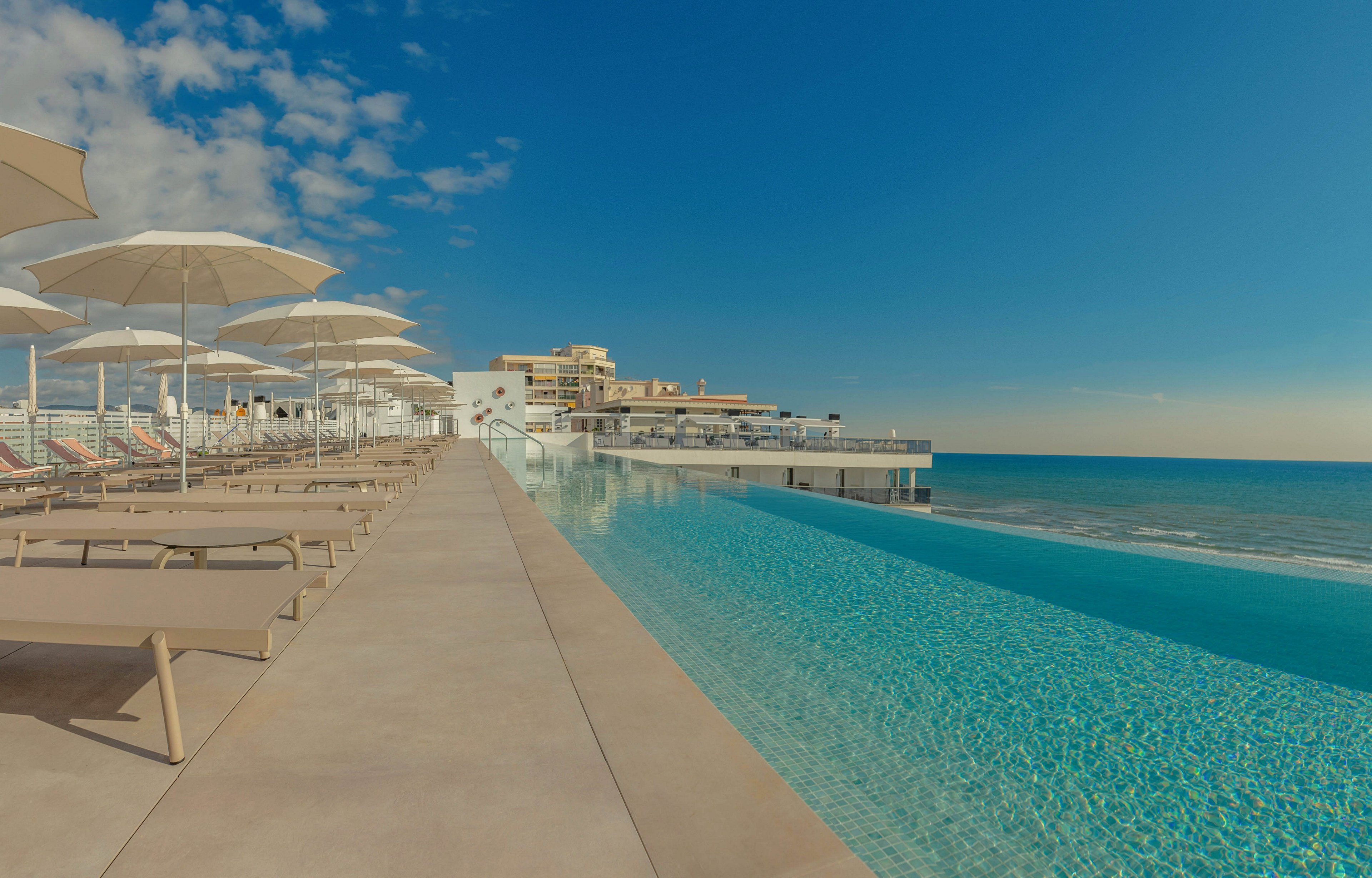 A long infinity pool stretches alongside a modern seaside resort, blending seamlessly into the calm ocean horizon. Empty lounge chairs and white umbrellas line the pool deck, suggesting a peaceful and uncrowded atmosphere. The sky is mostly clear with a few scattered clouds, enhancing the sense of openness and relaxation. Warm tones and soft lighting create a serene vacation mood.