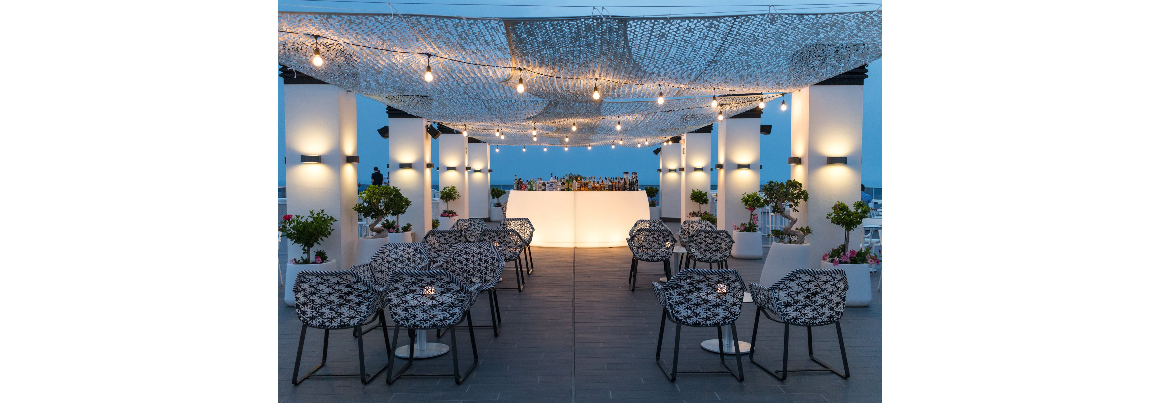 An elegant rooftop bar is arranged with patterned chairs facing a glowing white counter. Overhead, string lights and a mesh canopy create a cozy, intimate atmosphere as evening falls. Potted plants and flowers line the sides, adding greenery and color to the modern setting. The scene suggests a sophisticated outdoor venue for cocktails, events, or social gatherings by the sea.