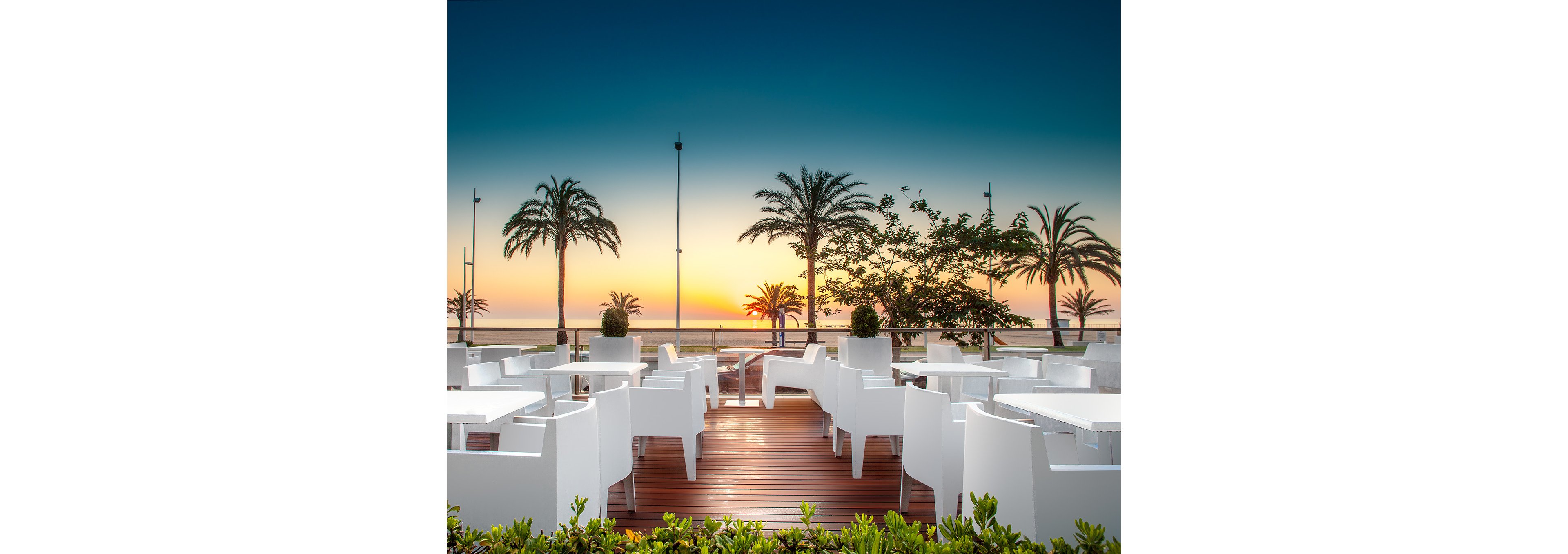 An empty outdoor restaurant terrace with sleek white tables and chairs overlooks the sea at sunset. Tall palm trees line the waterfront, silhouetted against a vivid gradient sky transitioning from deep blue to warm orange. The wooden deck and neatly trimmed greenery in the foreground add a polished, resort-like feel. The calm ocean and clear horizon create a tranquil, vacation atmosphere.