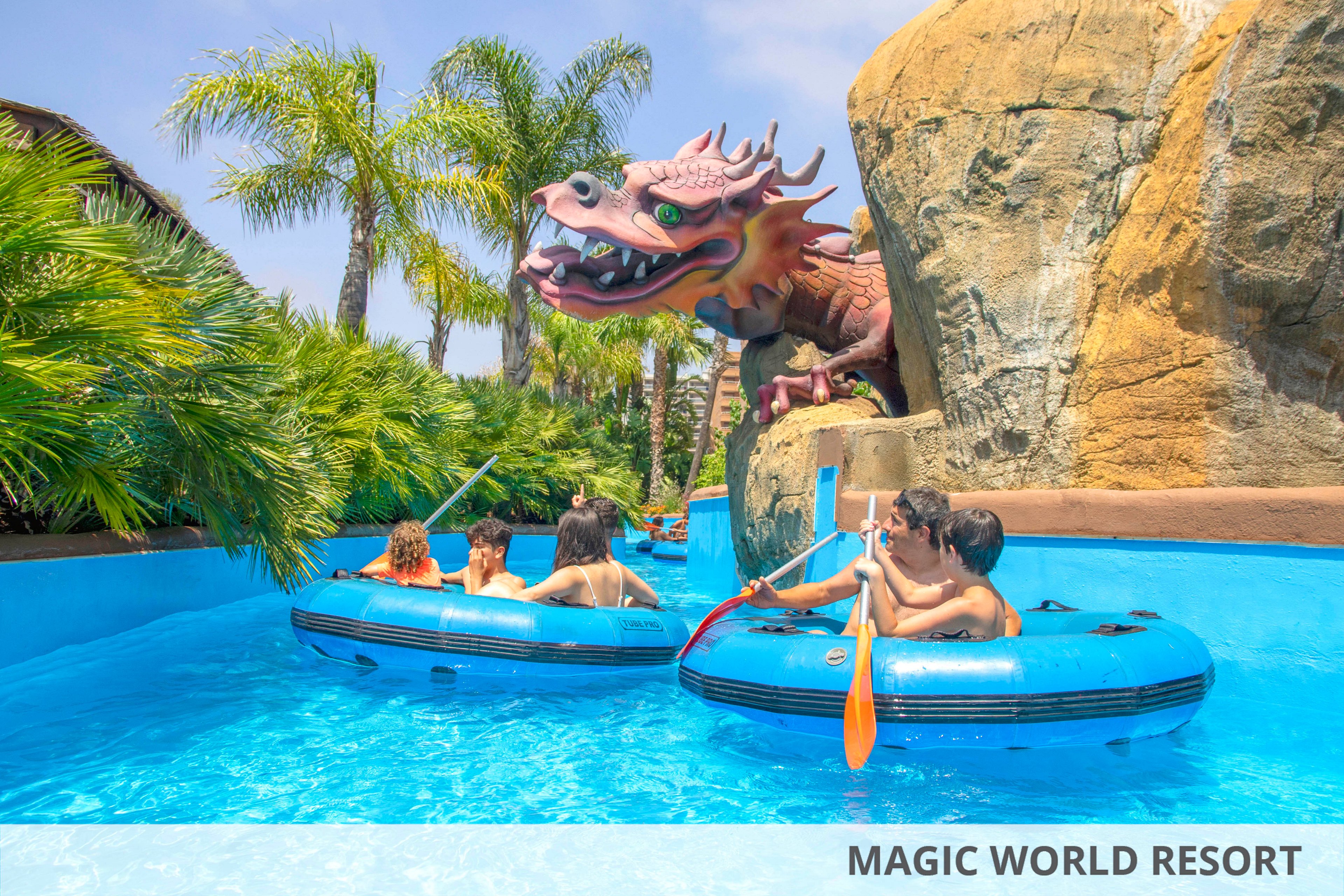 Several people float in blue inflatable rafts along a bright turquoise water channel at an outdoor amusement park. A large decorative dragon sculpture emerges from a rocky wall beside the ride, adding a fantasy theme to the scene. Lush green palm trees and tropical plants surround the attraction under a clear sunny sky. The overall atmosphere is fun, adventurous, and family-friendly.