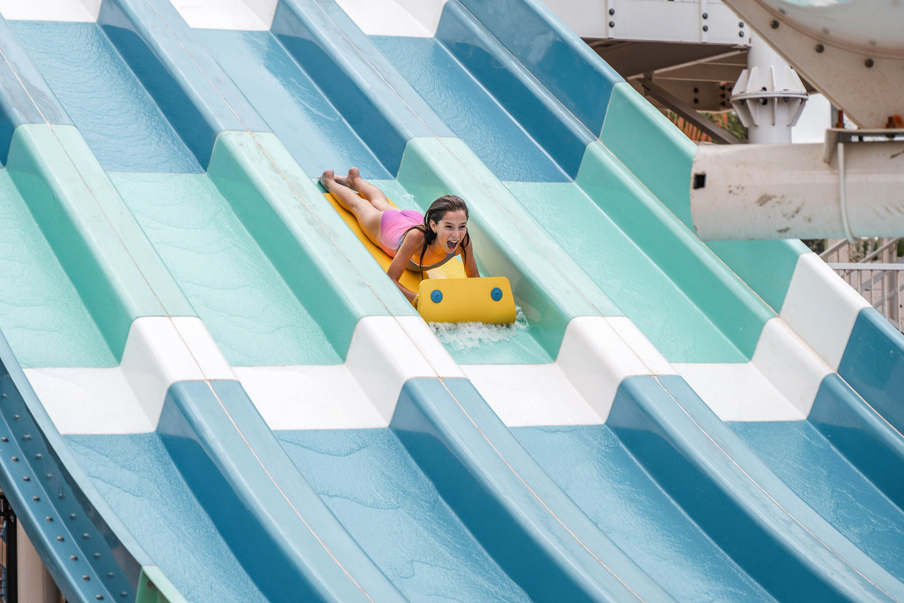 A young child rides a yellow mat down a multi-lane water slide at an outdoor water park. The slide features alternating turquoise and white lanes with water flowing beneath the rider. The child wears a pink swimsuit and is captured mid-descent, facing forward. Structural elements of the water park are visible in the background, emphasizing a fun recreational setting.