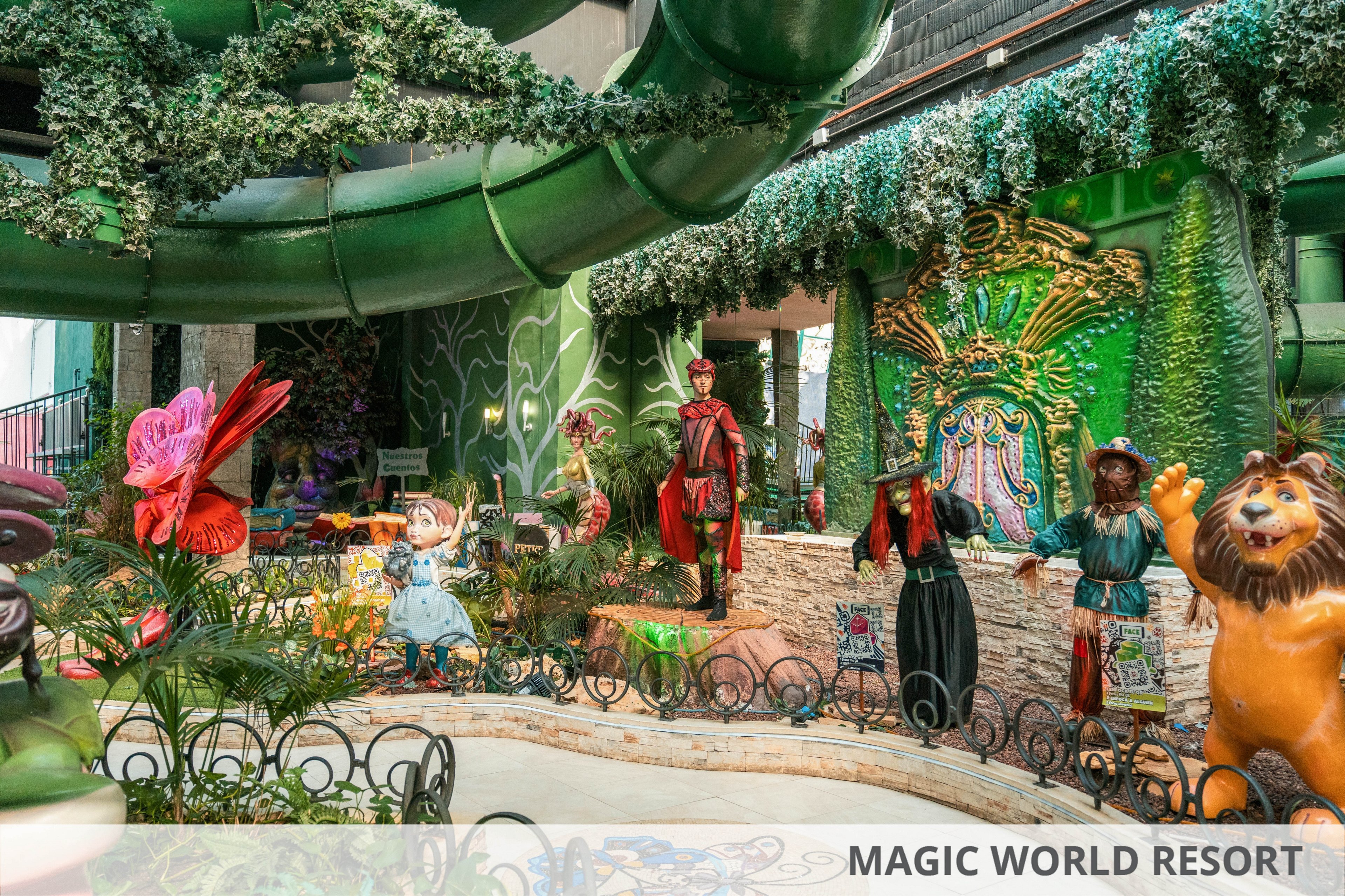 The image shows a vibrant fantasy-themed display with multiple storybook characters arranged in an indoor attraction. Figures of a girl, a lion, and other costumed characters stand among lush plants and decorative props. A large green slide structure curves overhead, while ornate green and gold backdrops create a magical atmosphere. The scene is filled with bright colors, whimsical sculptures, and detailed costumes that suggest a family-friendly entertainment setting.