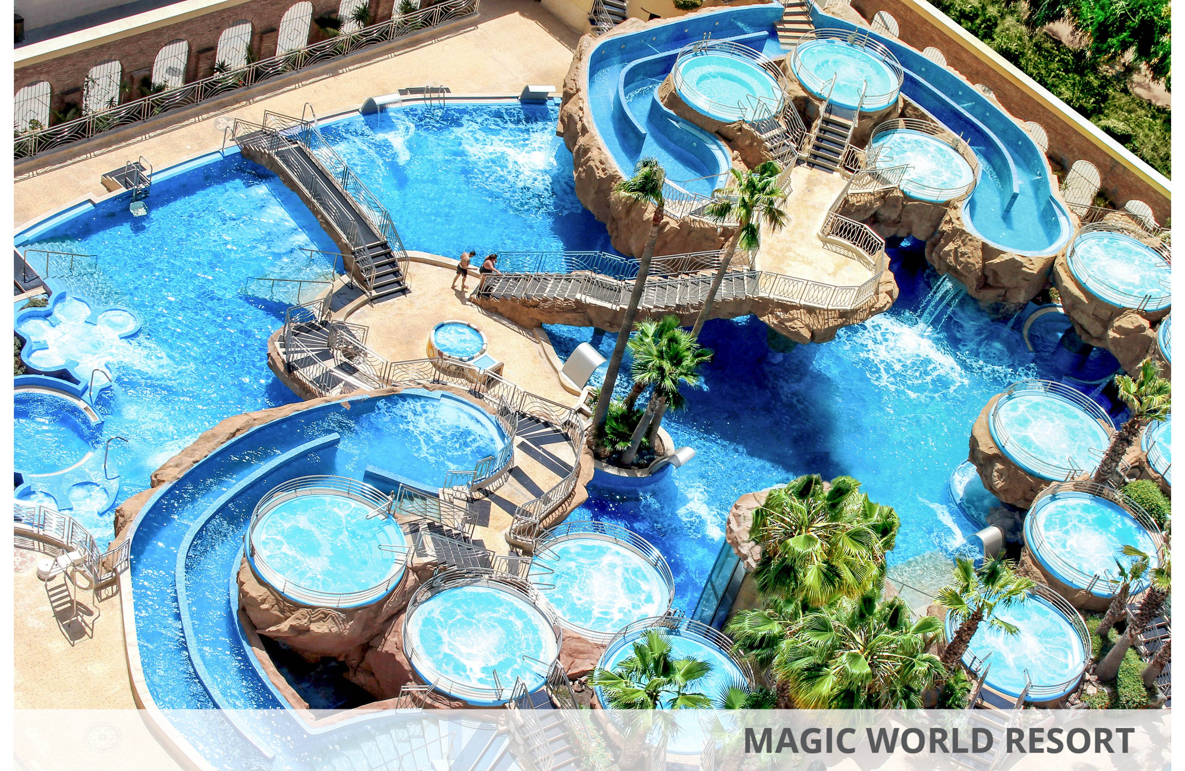 A large resort-style swimming pool complex features multiple interconnected pools and circular hot tubs. Elevated rock-like structures and stairways create different levels and lounging areas around the water. Palm trees and sunlit blue water give the scene a tropical vacation atmosphere. Two people relax near the center, emphasizing leisure and relaxation in this outdoor setting.