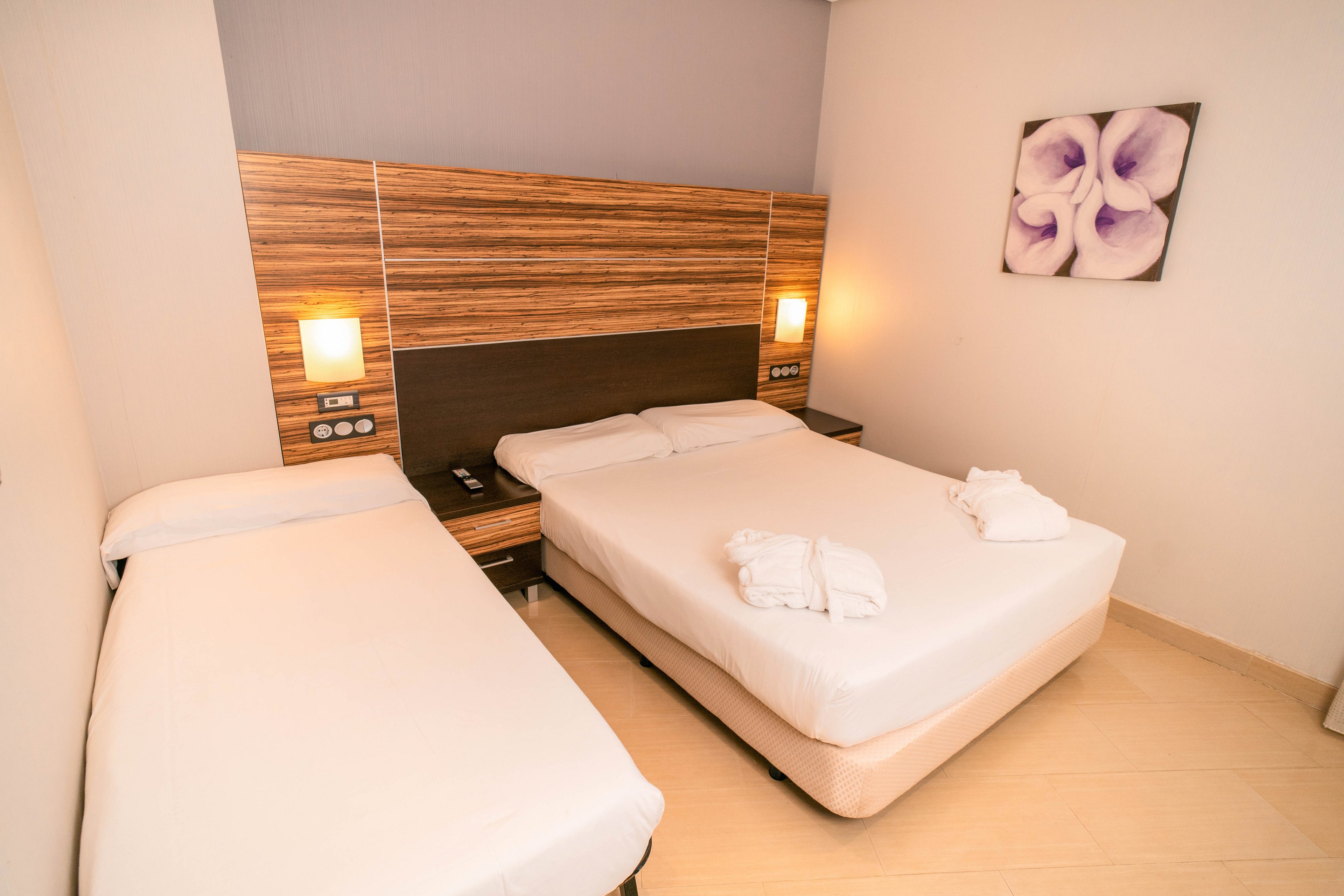 A contemporary hotel room features a double bed and a single bed with white linens and folded towels placed on the larger bed. The beds are set against a wooden headboard with built-in wall lamps providing warm ambient lighting. Light-colored flooring and neutral walls create a clean, minimalist atmosphere. A floral artwork hangs on the wall, adding a soft decorative touch to the space.