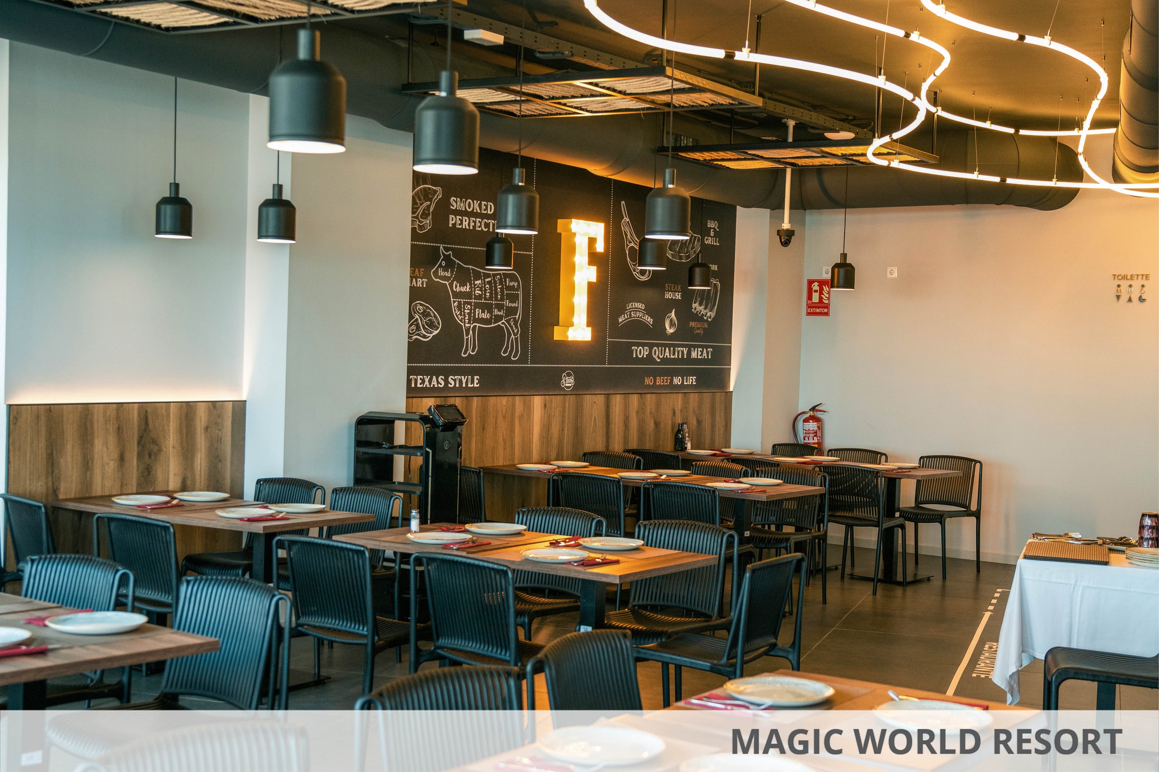 Contemporary restaurant dining area with neatly arranged tables and chairs is shown in warm ambient lighting. Plates and placemats are set on wooden tables, suggesting readiness for guests. A large black wall features illustrated meat cuts and text such as "SMOKED PROJECTS," "TOP QUALITY MEAT," and "TEXAS STYLE," along with a prominent letter "F." Overhead, curved ceiling lights and hanging pendant lamps add a stylish, modern touch to the space.