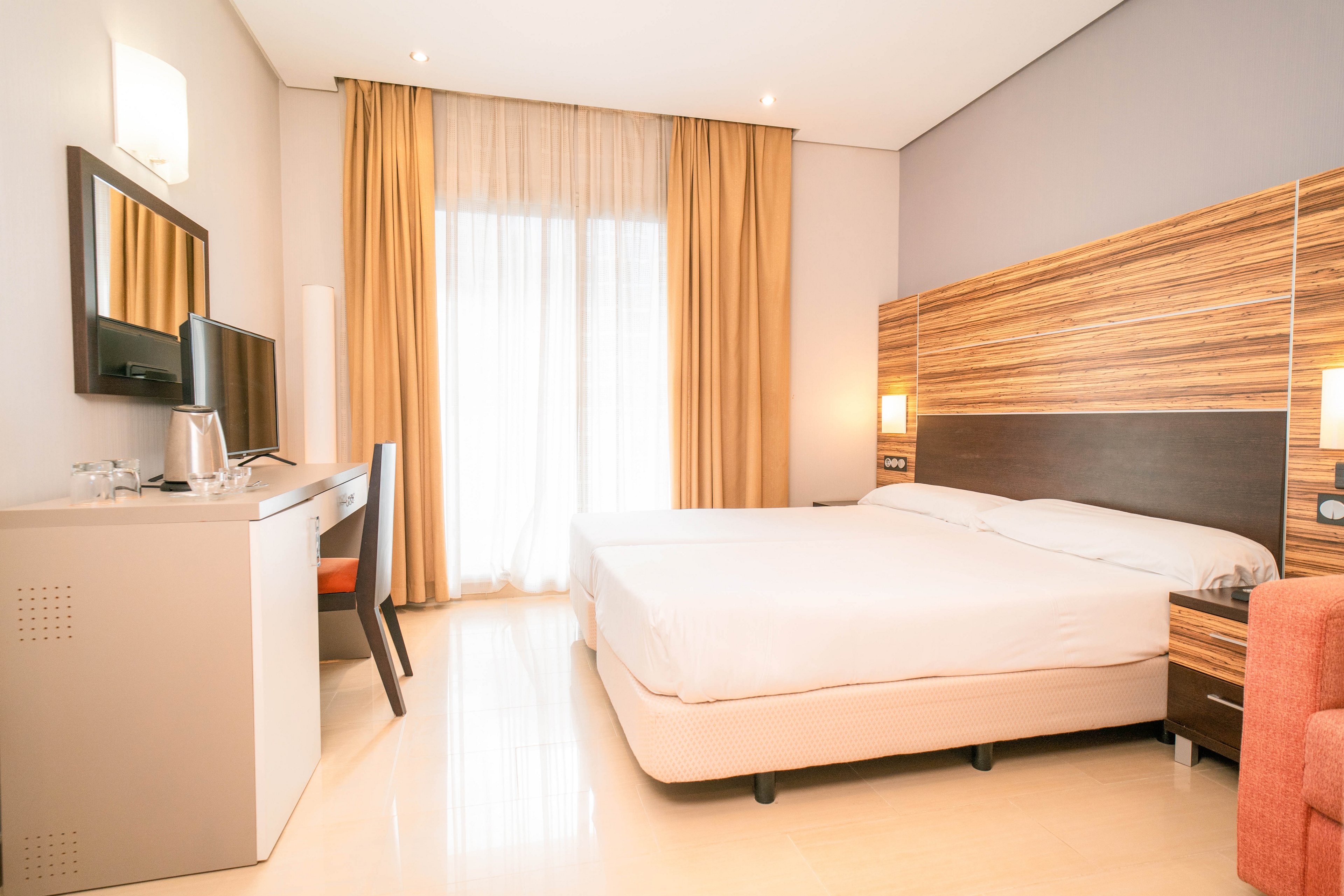 A contemporary hotel room features a large double bed with white linens and a wood-paneled headboard. A desk with a flat-screen TV, chair, and electric kettle sits opposite the bed. Floor-to-ceiling curtains cover a bright window or balcony door, allowing natural light to fill the space. Soft neutral tones and clean lines create a calm, minimalist atmosphere.