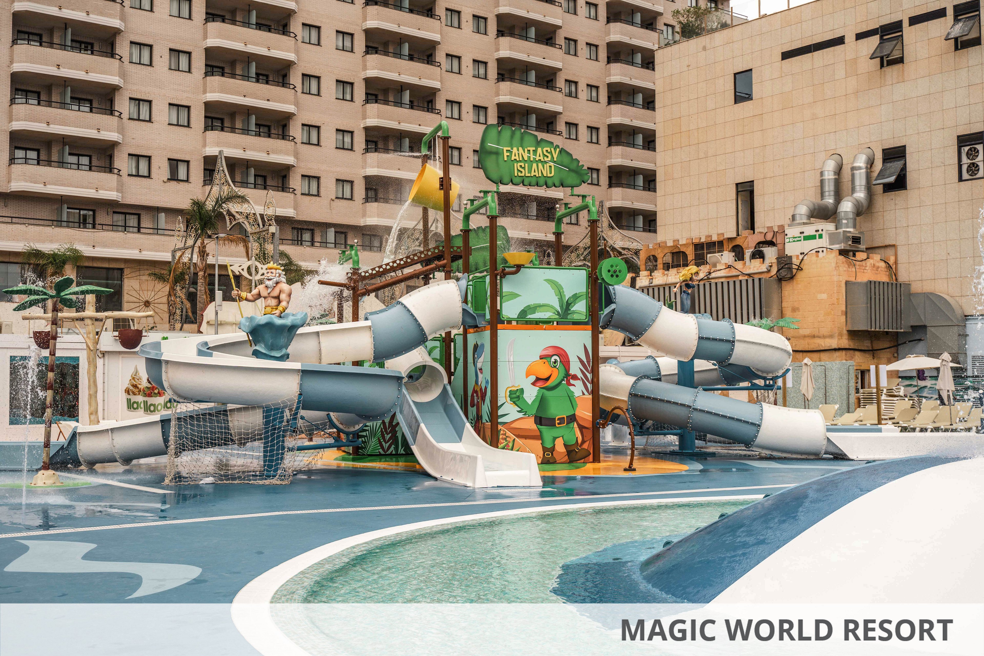 Colorful water play structure named Fantasy Island stands in an outdoor pool area. Multiple twisting slides and splash features are surrounded by cartoon tropical decorations and a parrot character. The playground is set against tall apartment buildings and a beige commercial facade, creating an urban resort atmosphere. Shallow pools and wet surfaces suggest a family-friendly water park environment.