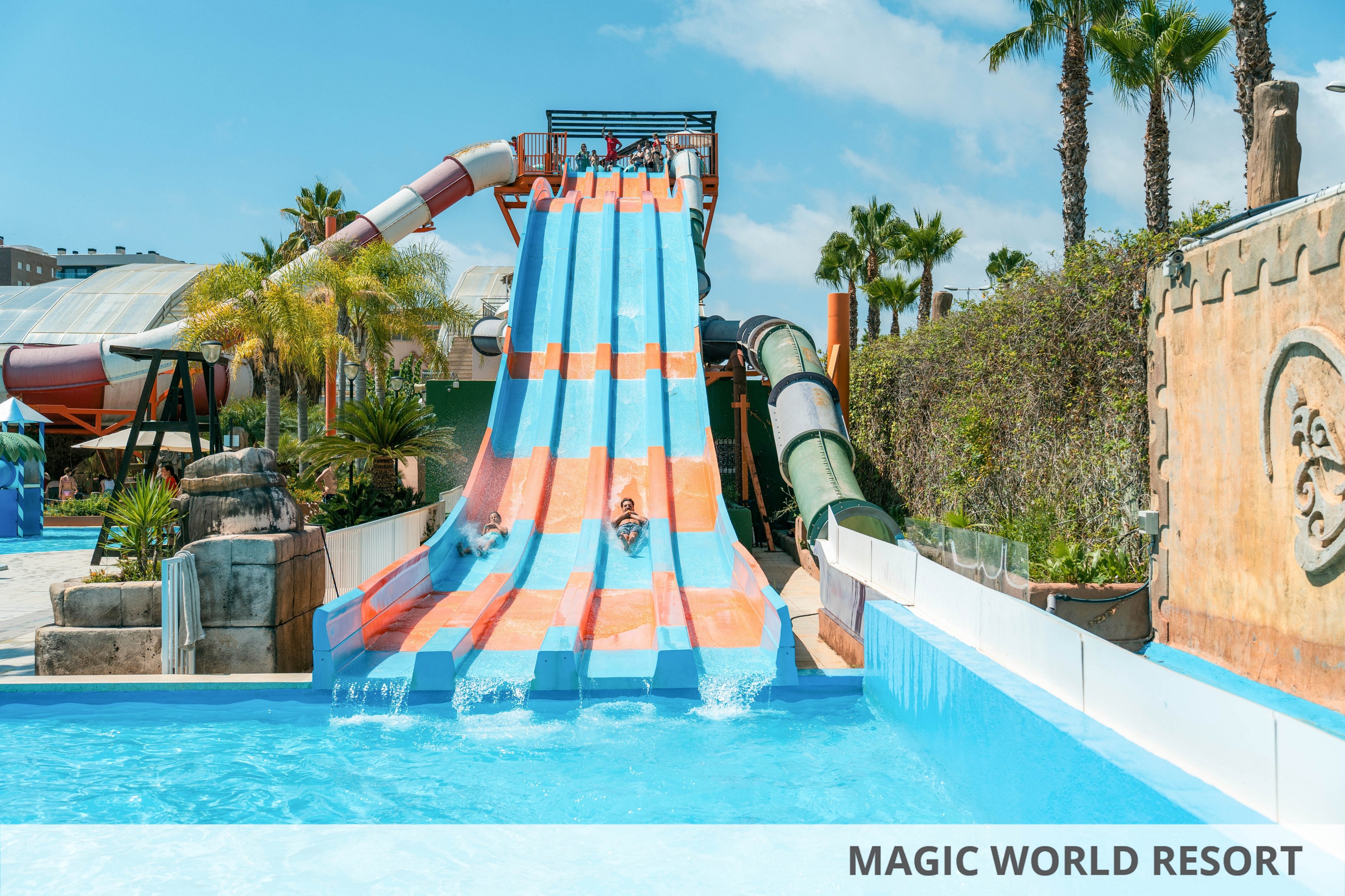 Several children slide down parallel blue and orange water slides into a bright blue pool. The scene is set in an outdoor water park with palm trees, greenery, and themed stone walls. Additional enclosed tube slides and park structures are visible in the background under a sunny sky. The overall atmosphere is fun, energetic, and summery.
