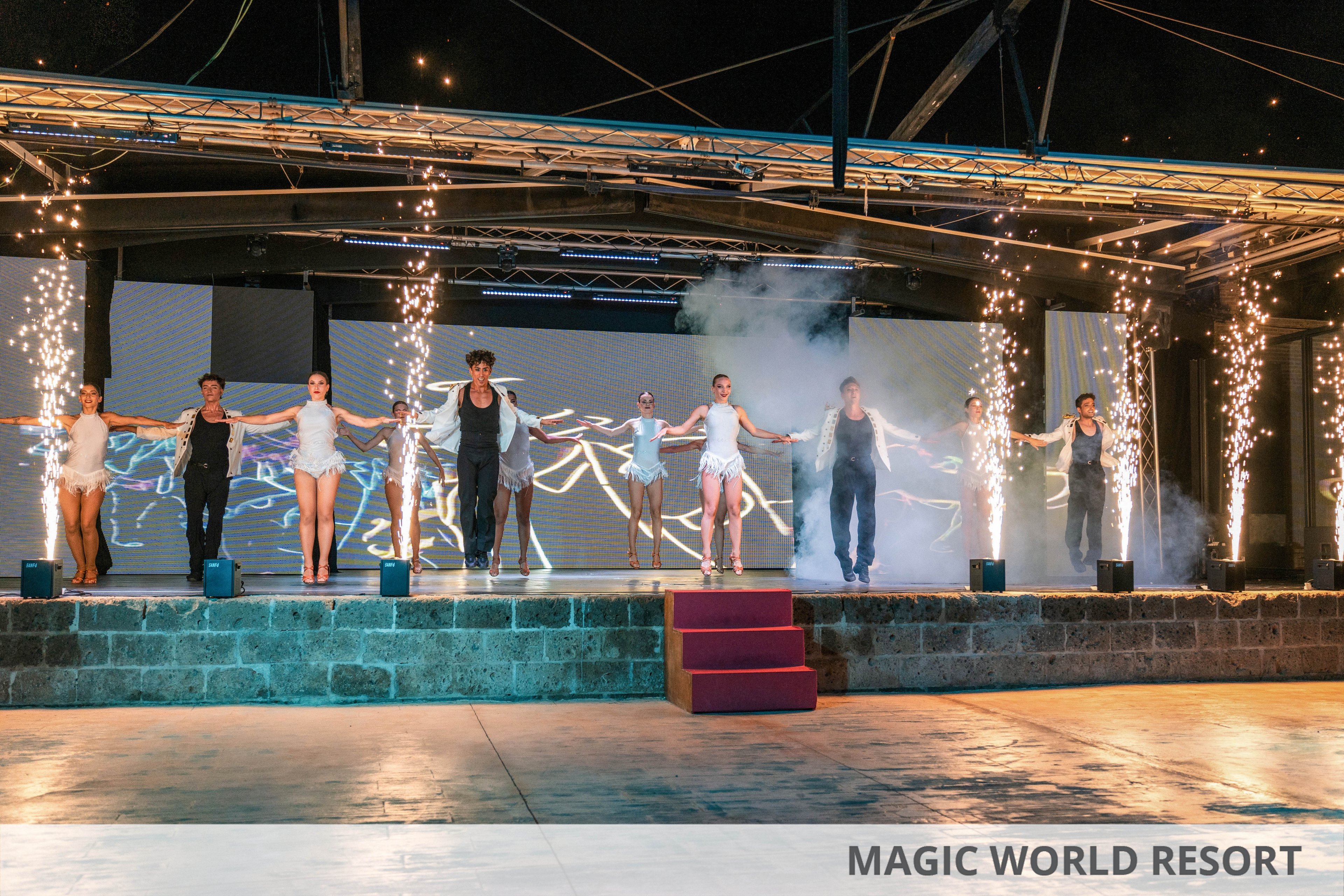 A group of dancers stands in a line on an outdoor stage, holding hands as they perform under bright pyrotechnic effects. Sparkling fireworks shoot upward from the front of the stage, framing the performers in dramatic light. Smoke and illuminated screens in the background add a dynamic, theatrical atmosphere. The scene suggests a choreographed show or event with a festive, energetic mood.
