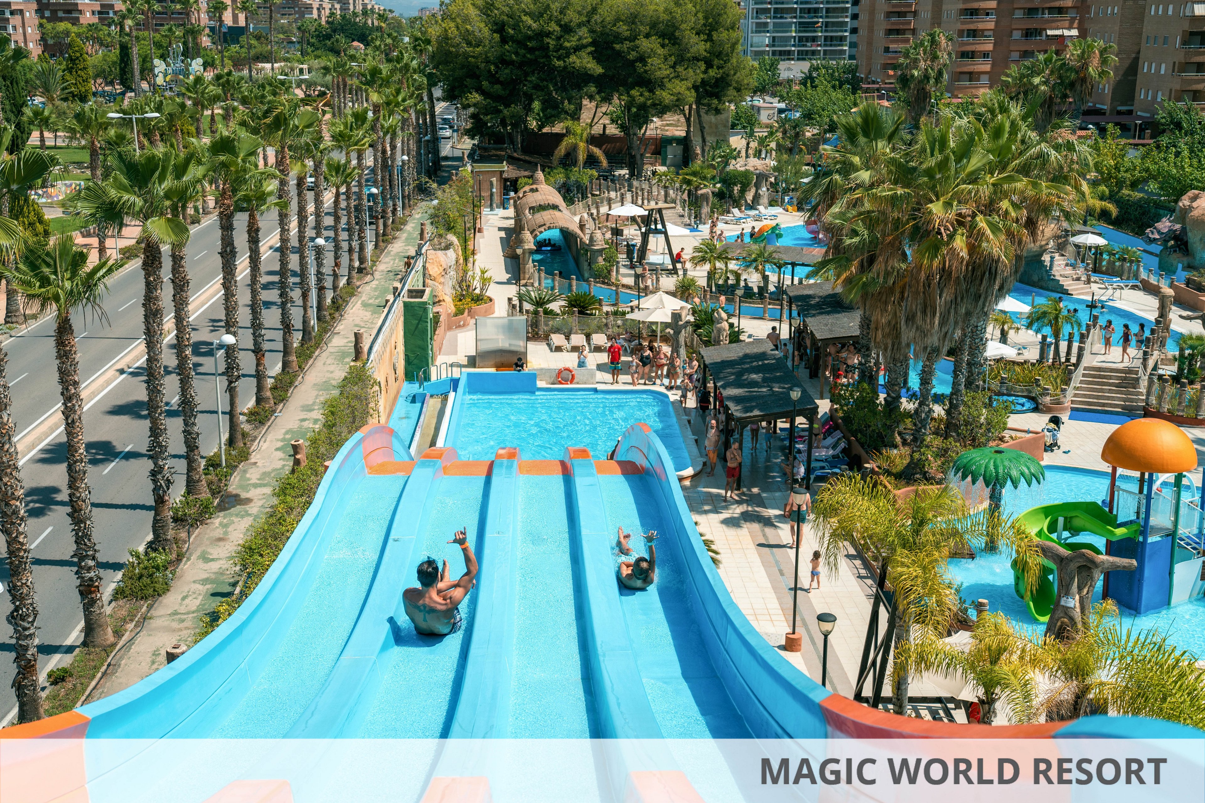 The image shows several people riding down wide blue water slides at a busy outdoor water park. Palm trees line the area, and multiple pools, play structures, and shaded seating are visible around the slides. Colorful features such as orange and green water play equipment add to the vibrant, summery atmosphere. In the background, urban buildings and a road with palm trees frame the recreational setting.