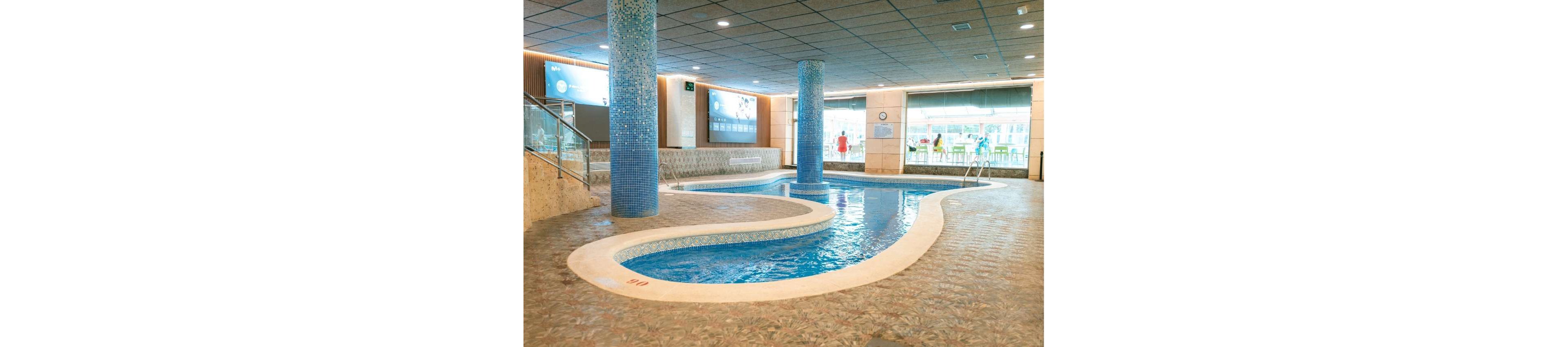 An indoor spa-style pool with a winding, curved shape is the main focus of this image. The pool is surrounded by tiled flooring and features blue mosaic-tiled columns rising from the water. Large windows and illuminated wall displays brighten the modern interior space. A few people can be seen in the background near the far end of the pool area.