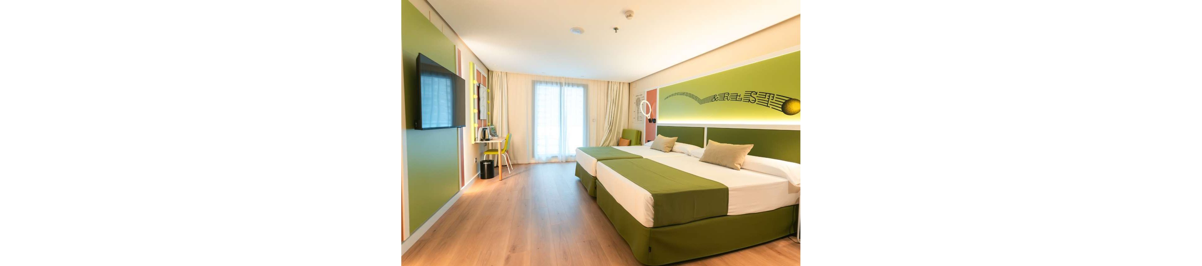 A contemporary hotel room features two large twin beds with green bedskirts and beige pillows. The space is decorated in fresh green and white tones with a wall-mounted flat-screen TV opposite the beds. A desk and chair sit near the window, which is covered by sheer curtains allowing natural light to fill the room. Wooden flooring and minimalist decor create a clean, inviting atmosphere suitable for travelers or vacation stays.
