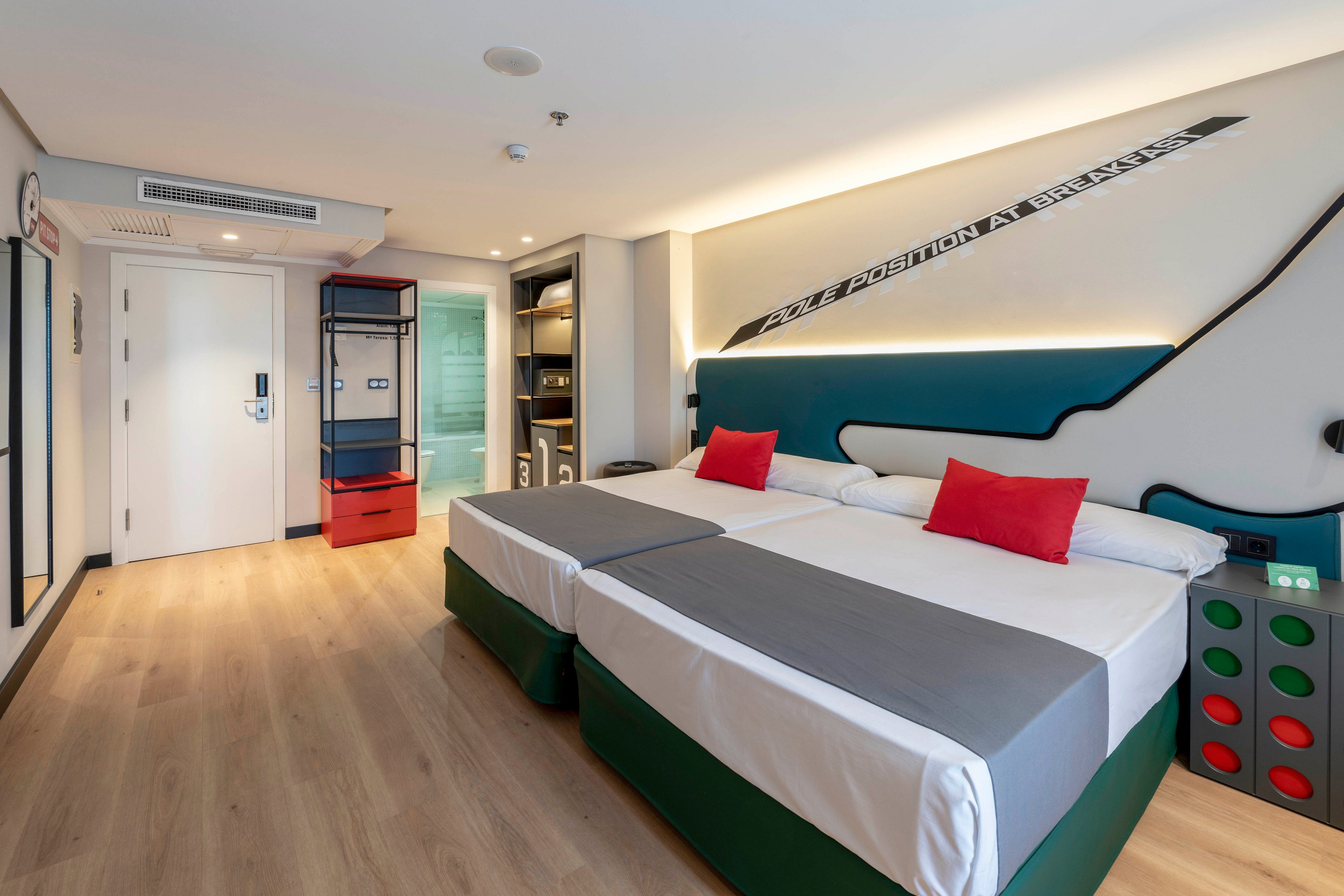 A contemporary hotel room features two neatly made twin beds with gray runners and red accent pillows. The space has light wooden flooring, a minimalist headboard design, and playful decor including a large dice-shaped bedside table. Built-in shelving, a wardrobe, and a glass-walled bathroom area are visible near the entrance. Soft ambient lighting and clean lines create a bright, welcoming atmosphere suitable for travelers.