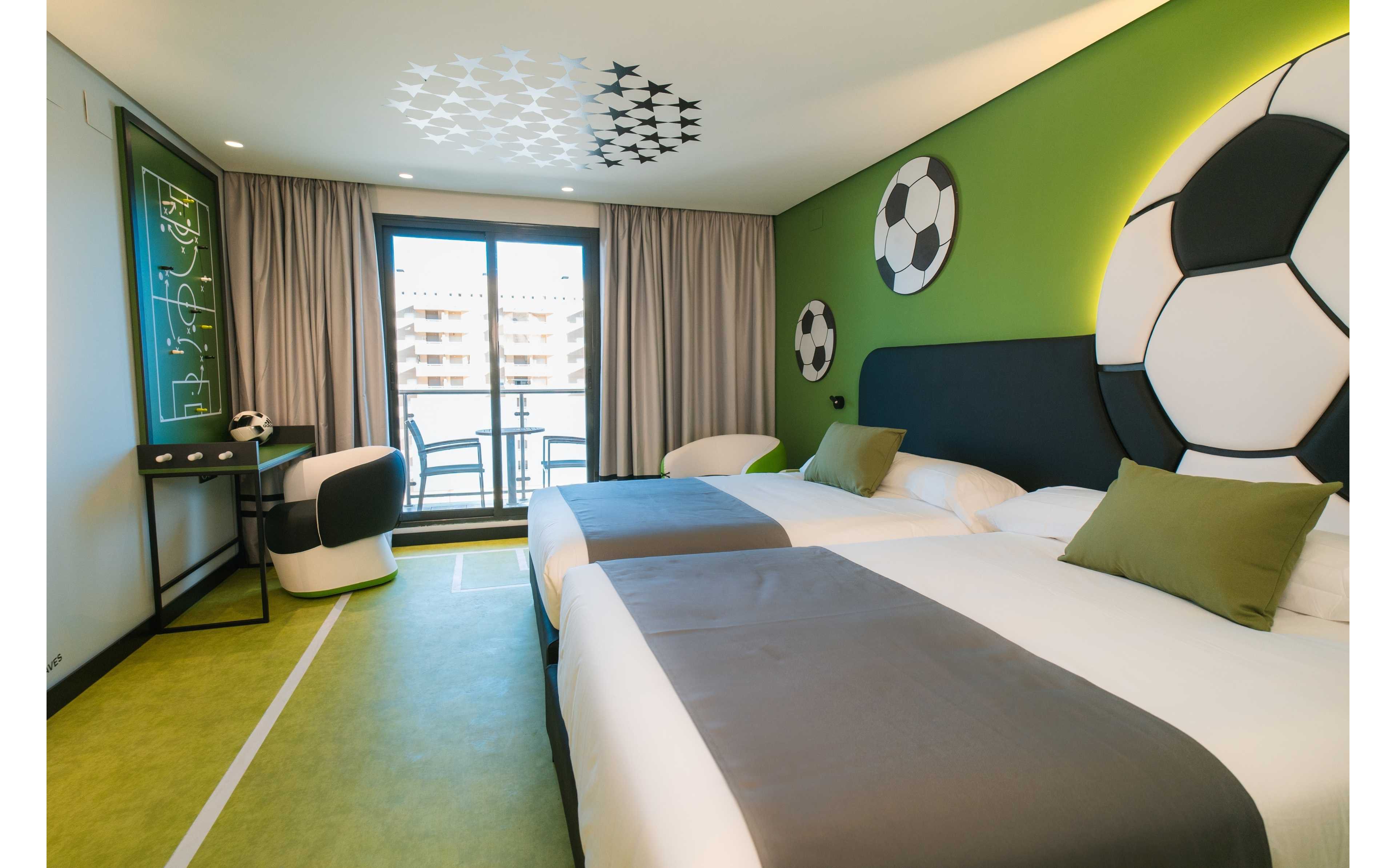 Contemporary hotel room decorated with a soccer theme features two neatly made beds with gray runners and green accent pillows. The walls and floor incorporate green field-inspired elements, including large soccer ball graphics and a painted pitch line. A desk area with a soccer-patterned chair and a tactical chalkboard-style artwork sits near the window. Floor-to-ceiling curtains frame a glass door leading to a balcony with an urban view, creating a bright and playful atmosphere.