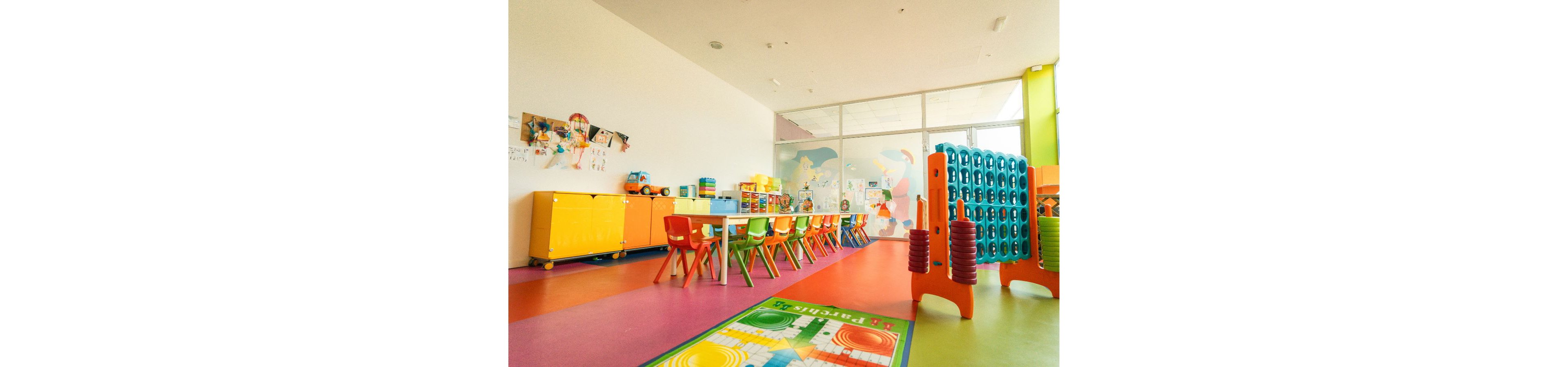 A bright preschool classroom features a long table lined with small colorful chairs. The room includes storage cabinets, building blocks, and various toys neatly arranged along the walls. A soft play structure and a vibrant floor mat add to the playful environment. Large windows and wall murals create an open, cheerful atmosphere designed for young children.