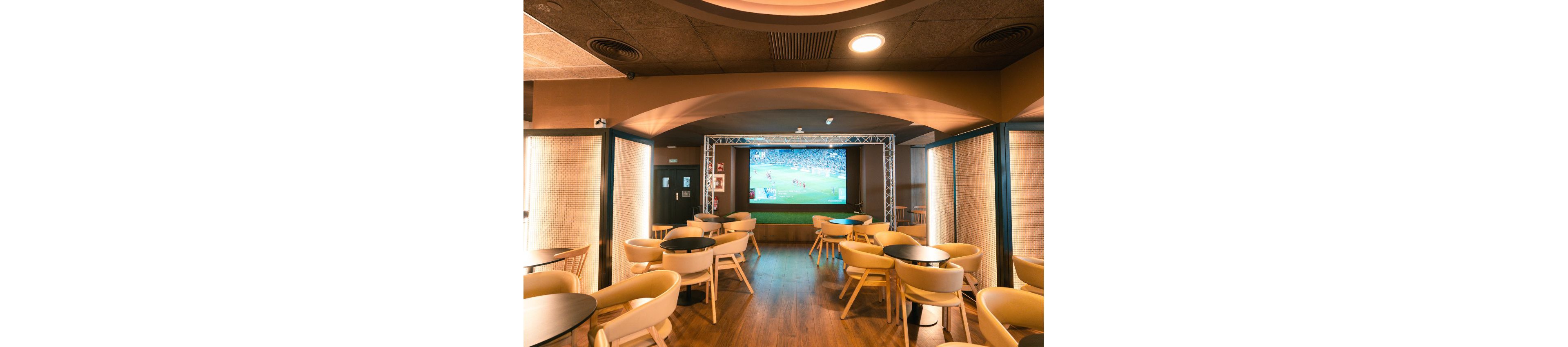 A contemporary lounge bar features multiple round tables and light-colored chairs arranged on a wooden floor. At the far end of the room, a large projection screen displays a live soccer match. Warm ambient lighting and arched ceiling details create a cozy, inviting atmosphere. The space appears ready for guests to watch sports events in a relaxed social setting.