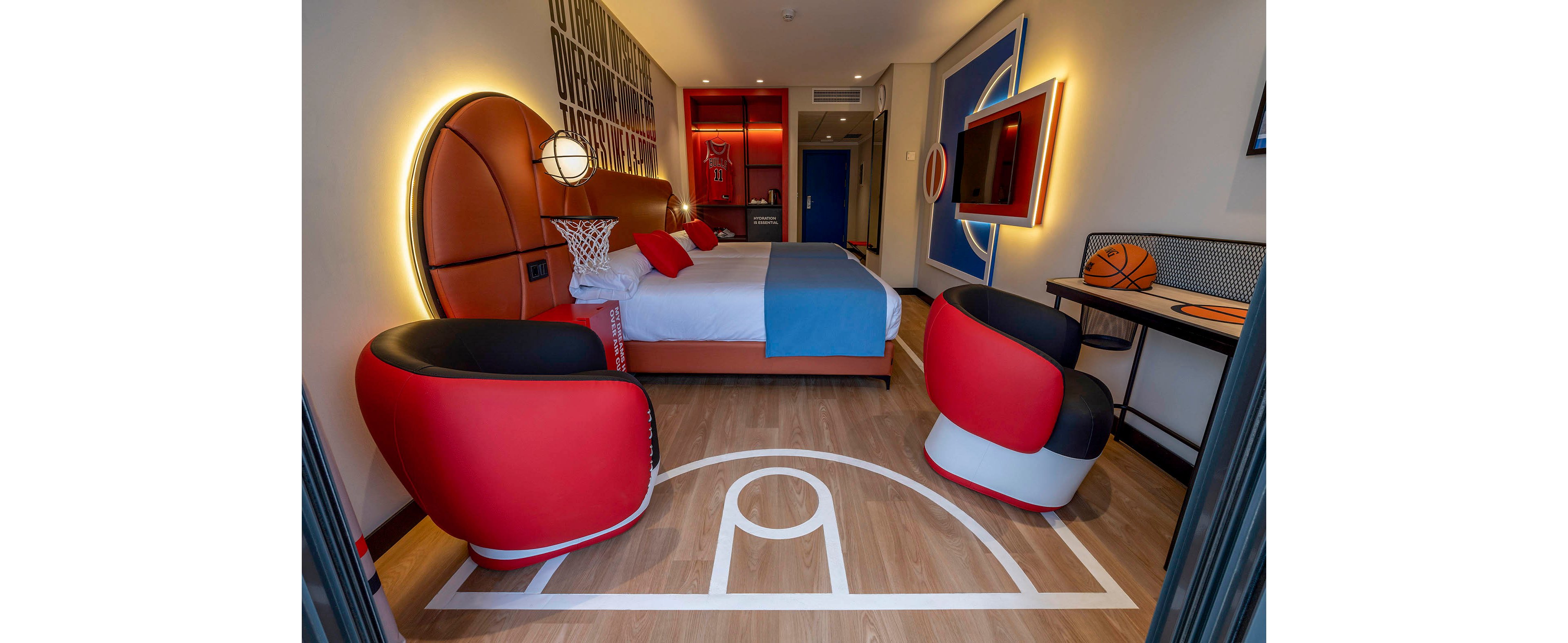 A modern hotel room is decorated with a bold basketball theme. The bed headboard resembles a giant basketball and a small hoop is mounted at the foot of the bed. Two red and black basketball-inspired armchairs face a desk with a basketball on top. Court lines are painted on the wooden floor, and wall art continues the sports motif with bright lighting accents.
